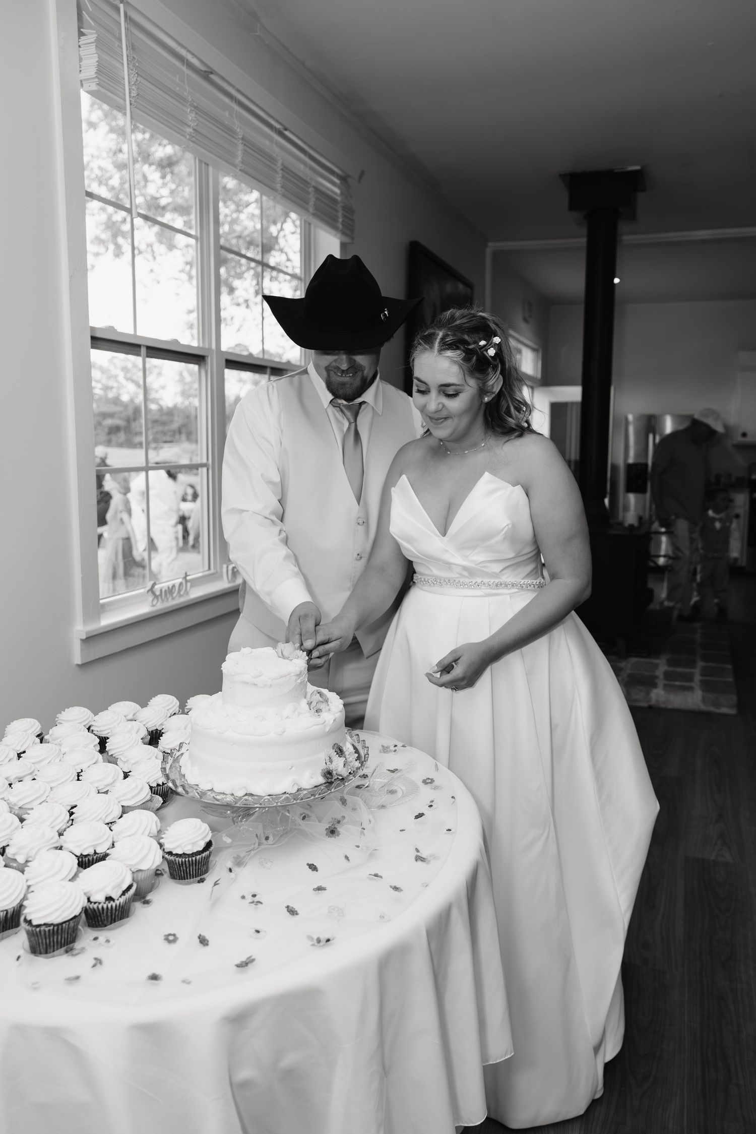 Bride and groom cutting wedding cake in black and white photo, groom wearing cowboy hat, surrounded by cupcakes.
