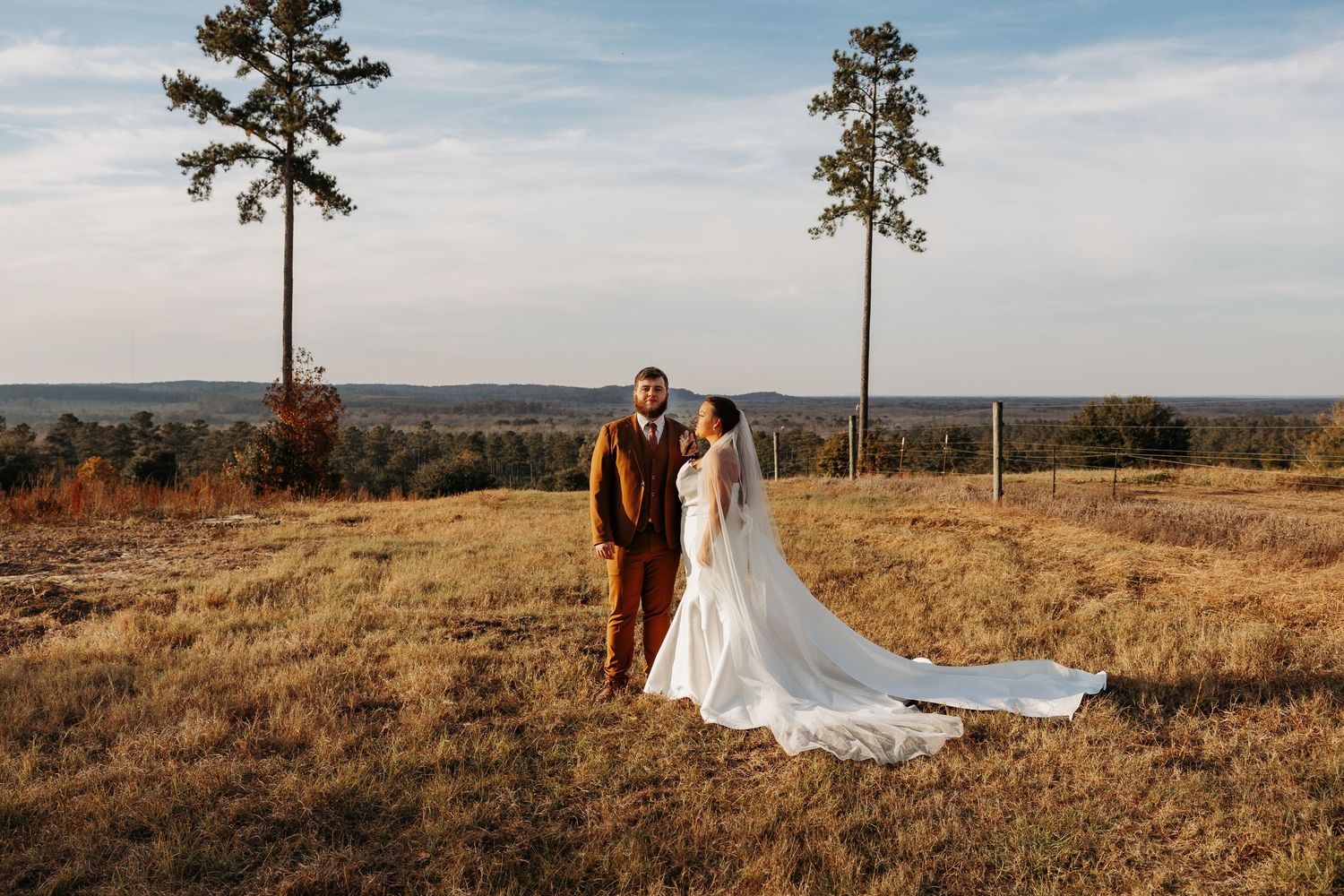 Bride and groom portrait in golden field with tall pine trees and open sky at outdoor wedding.