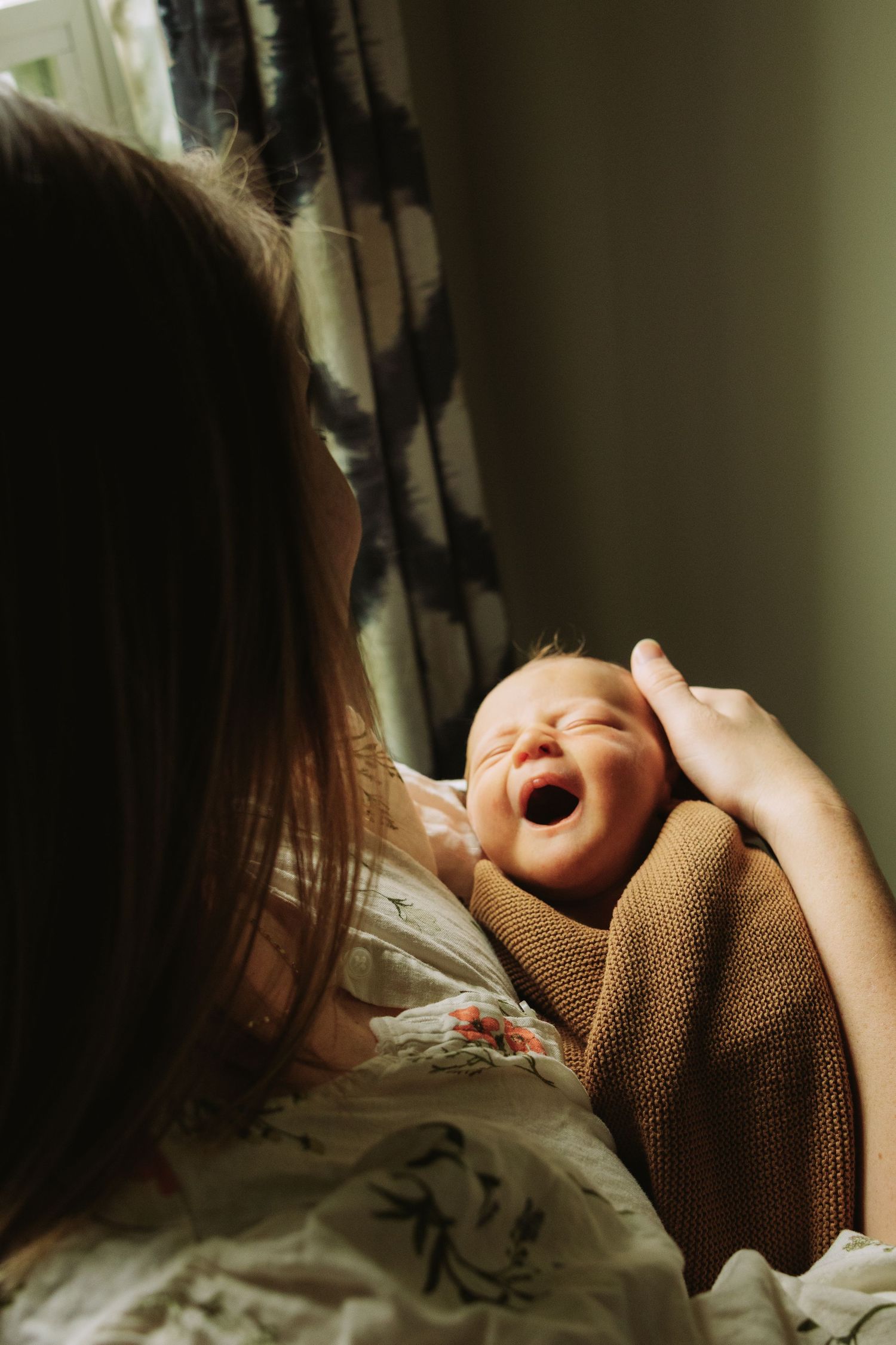 A joyful newborn baby wrapped in brown blanket laughing while being held near a window with curtains.