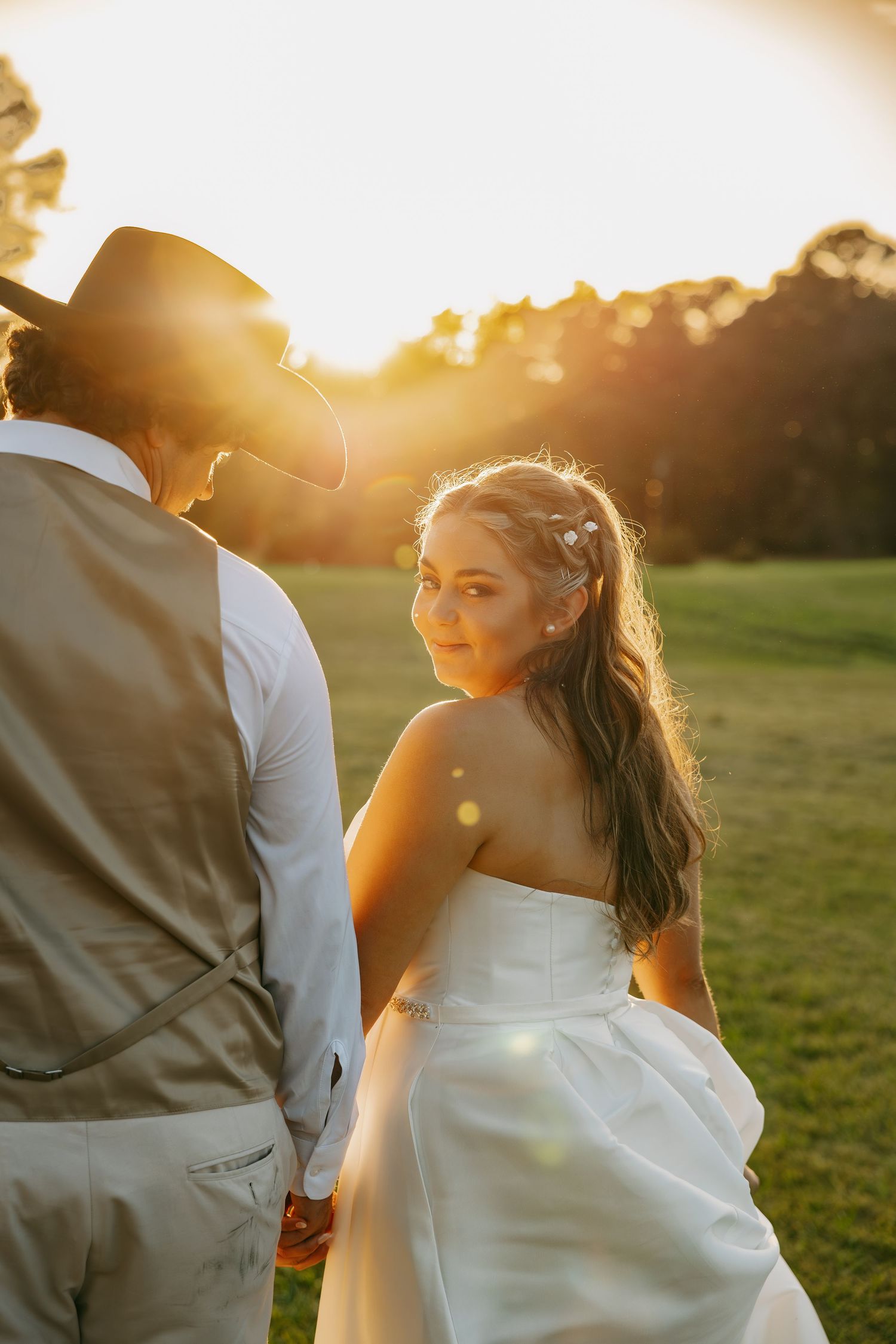 Bride in white strapless gown looks back smiling, holding hands with groom in vest and cowboy hat at golden sunset.