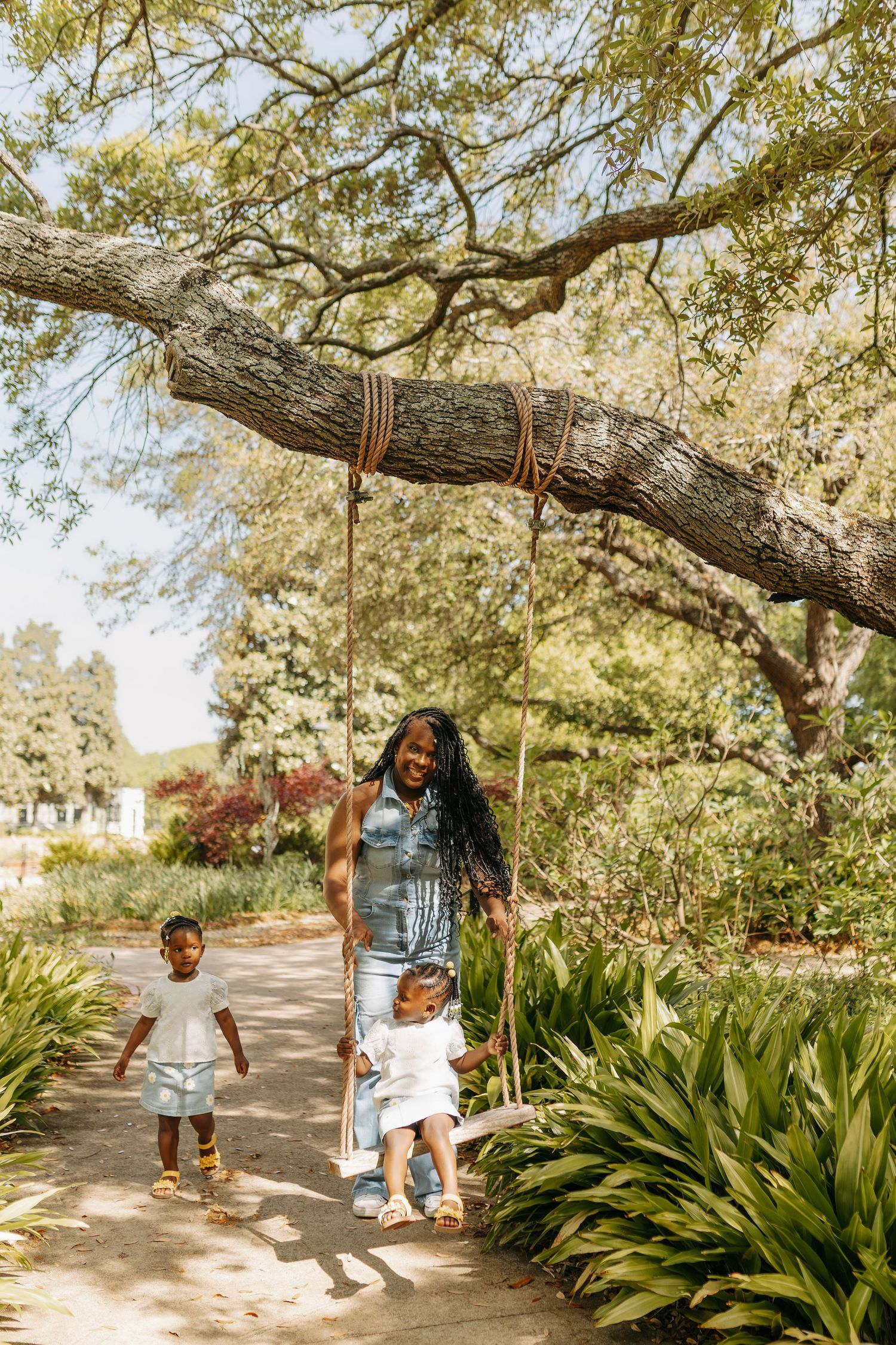 A mother with braids pushes her toddler on a rope tree swing while another child walks nearby in a sunny park.