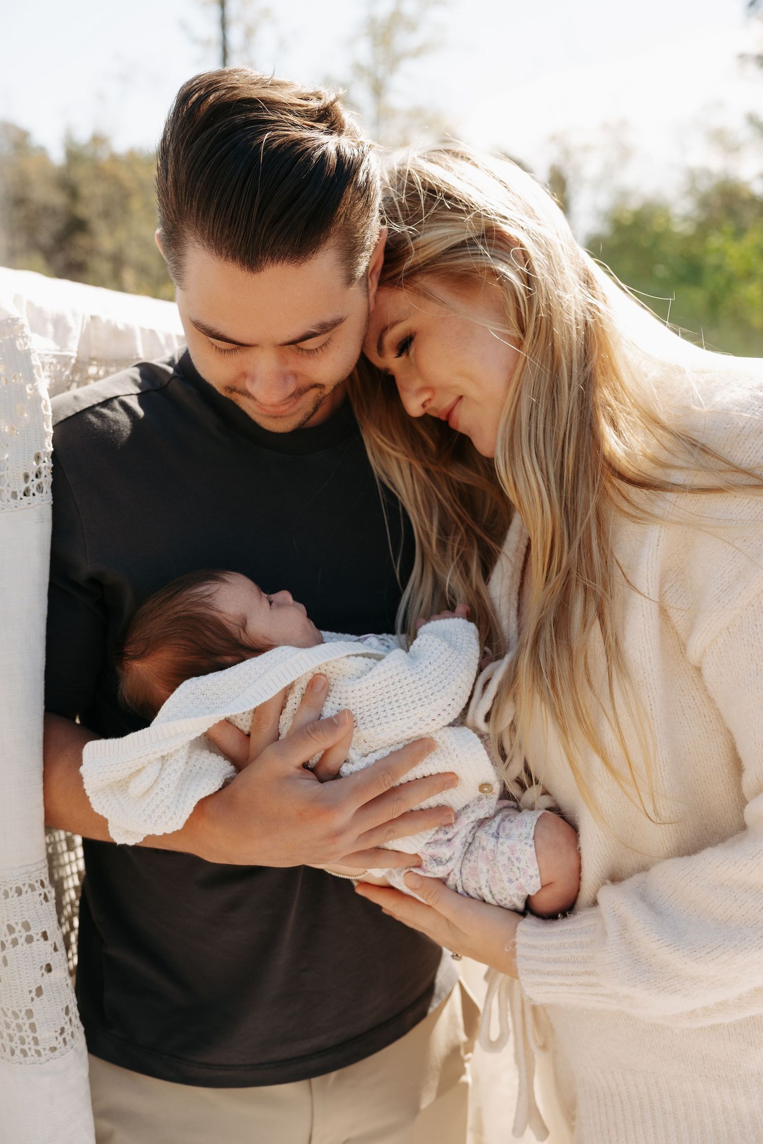 Young couple with dark and blonde hair tenderly holding a swaddled newborn baby outdoors in warm sunlight.