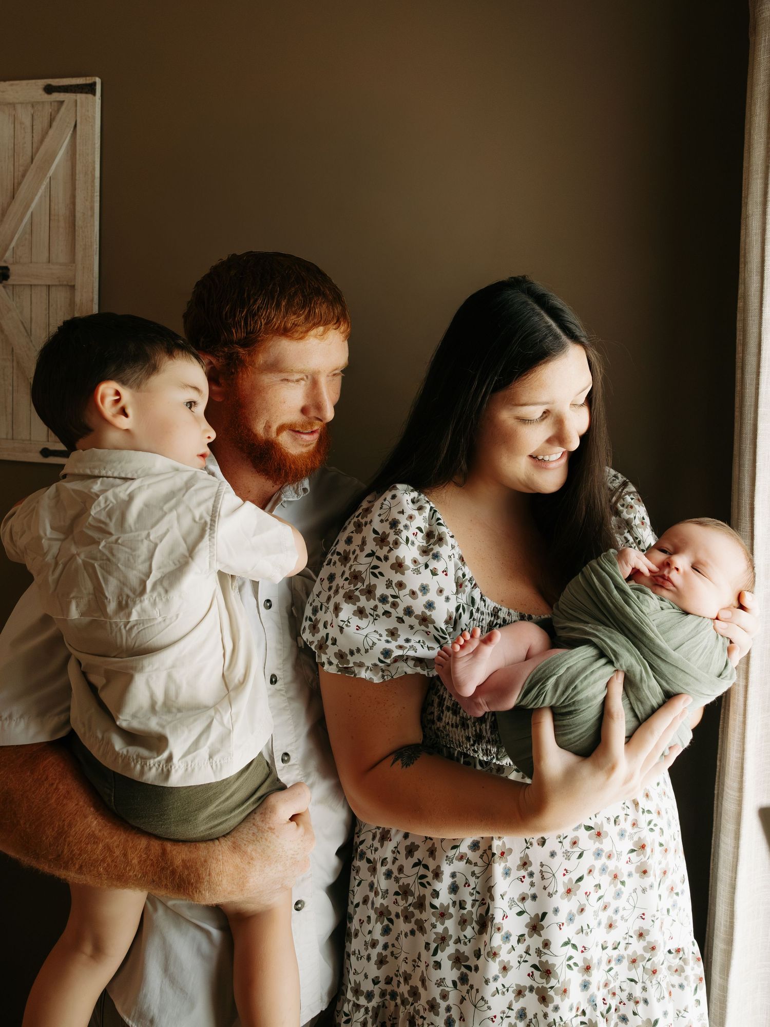 A young family with a redheaded father holding a toddler and a dark-haired mother cradling a newborn baby.