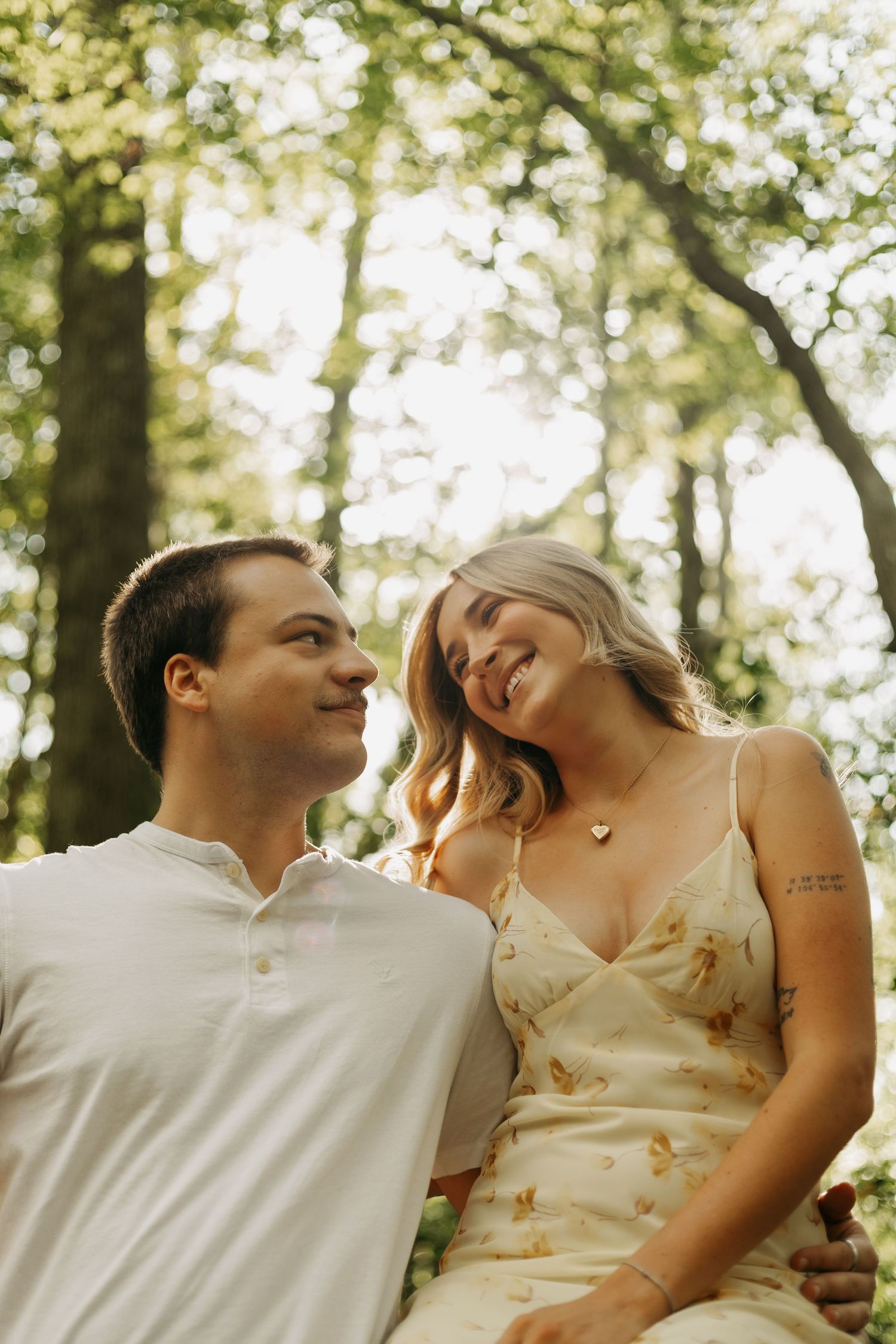 A smiling couple sitting together outdoors in a sunlit forest, woman in floral dress leaning toward man in white shirt.