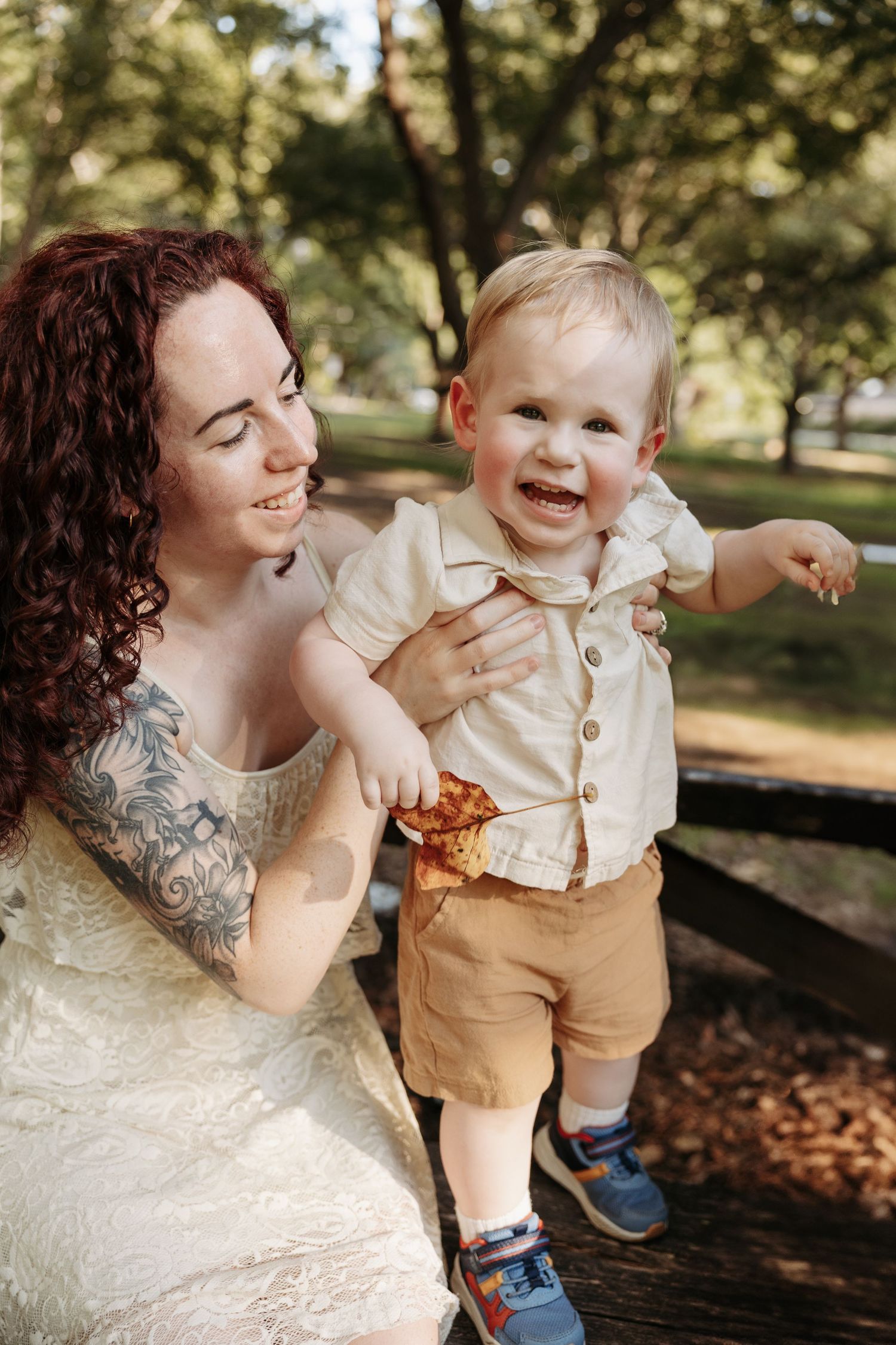 A smiling redhead tattooed woman in a white lace dress holds a laughing toddler boy in a sunny park setting.
