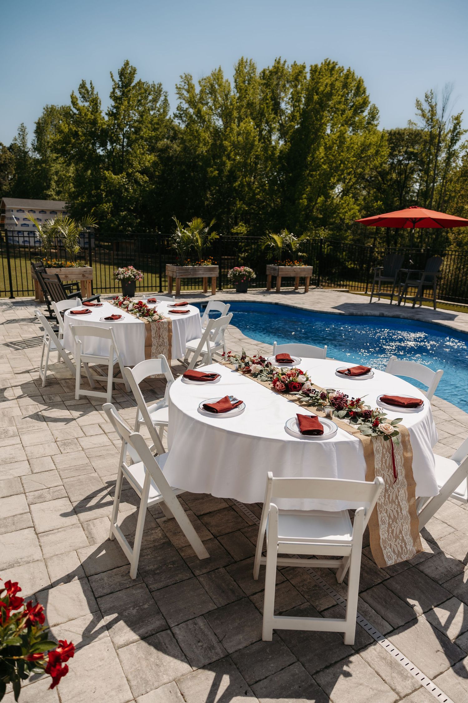 Elegant poolside wedding reception tables with white linens, red napkins, and floral centerpieces under blue sky.