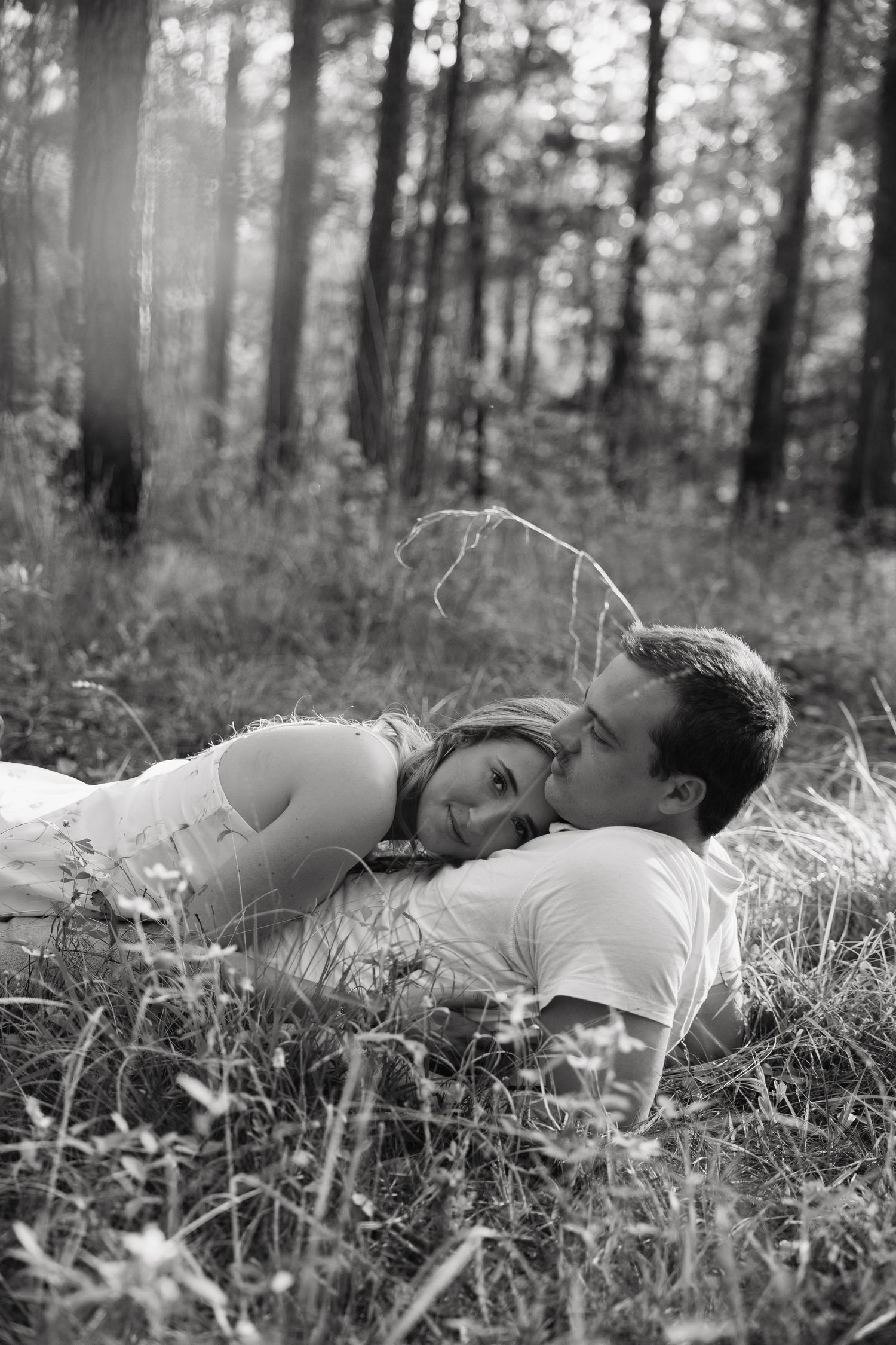Black and white photo of a romantic couple lying in a forest meadow, man kissing woman on cheek as she smiles.
