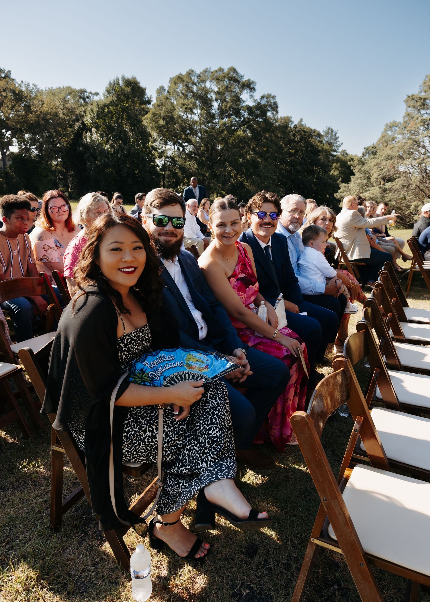 Wedding guests seated in wooden chairs at outdoor ceremony smile at camera on sunny day.