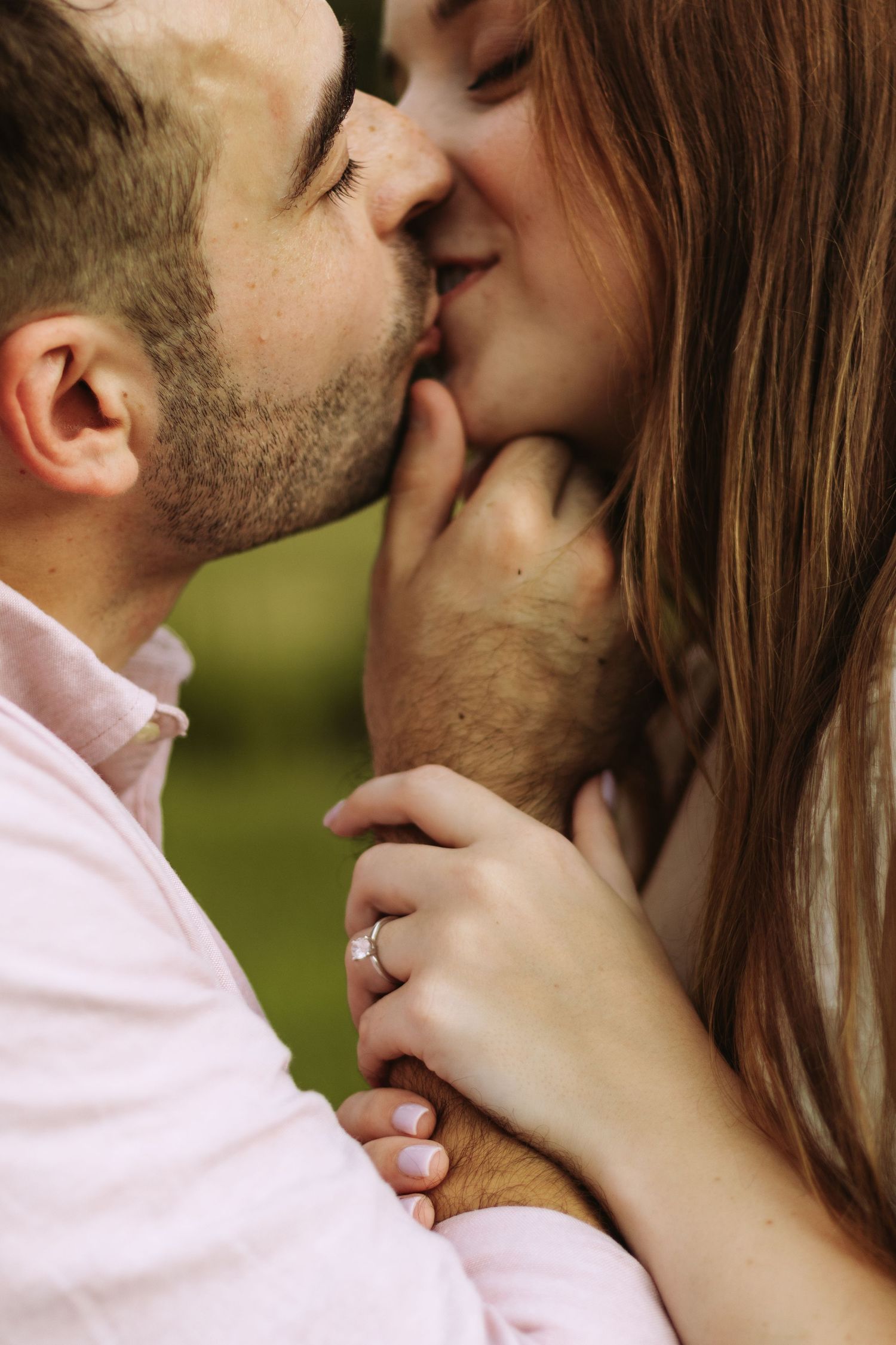Couple sharing a romantic kiss outdoors, woman wearing an engagement ring, man in pink shirt, green background.