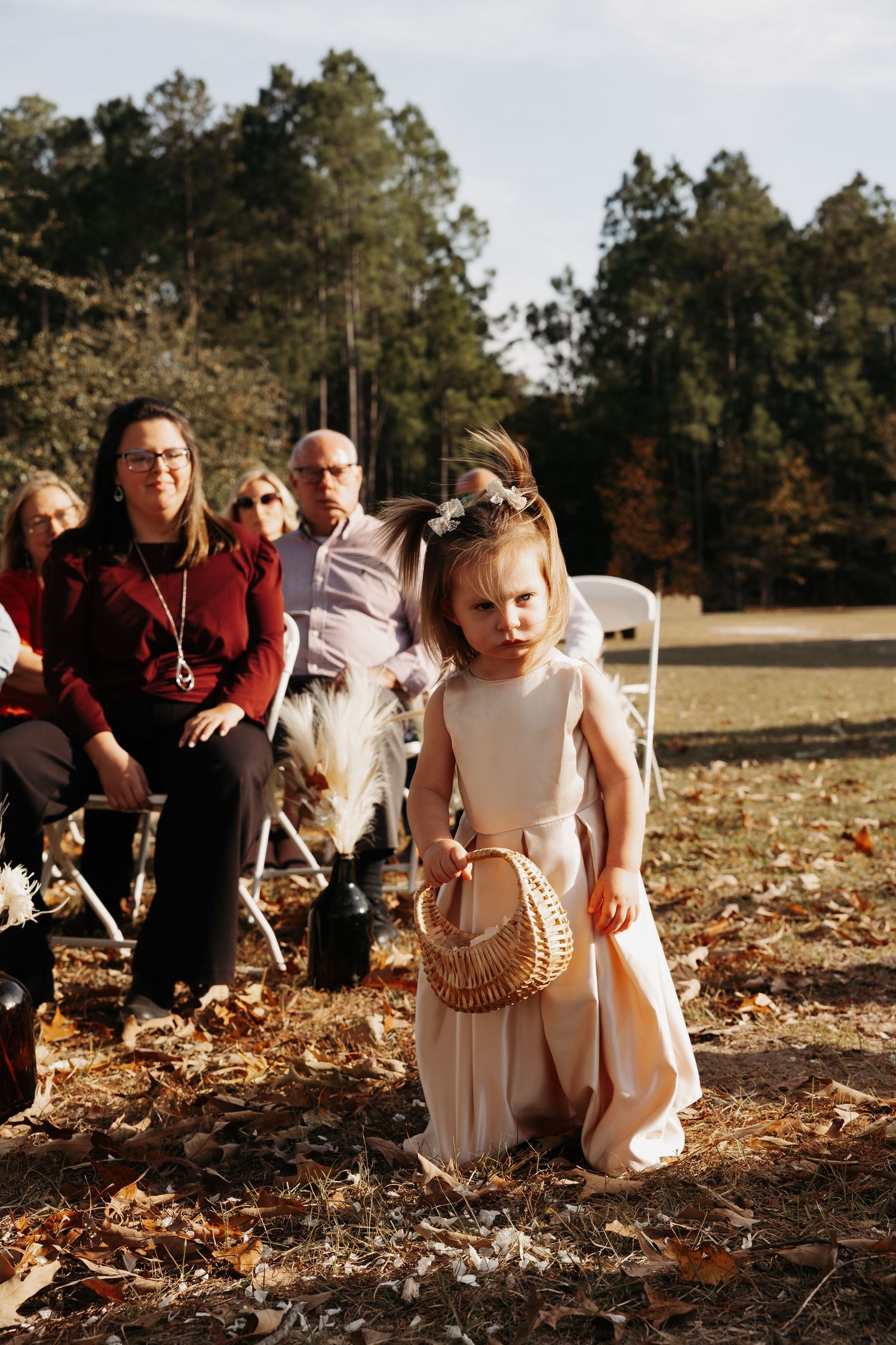 Flower girl in blush dress carrying basket down outdoor fall wedding aisle.