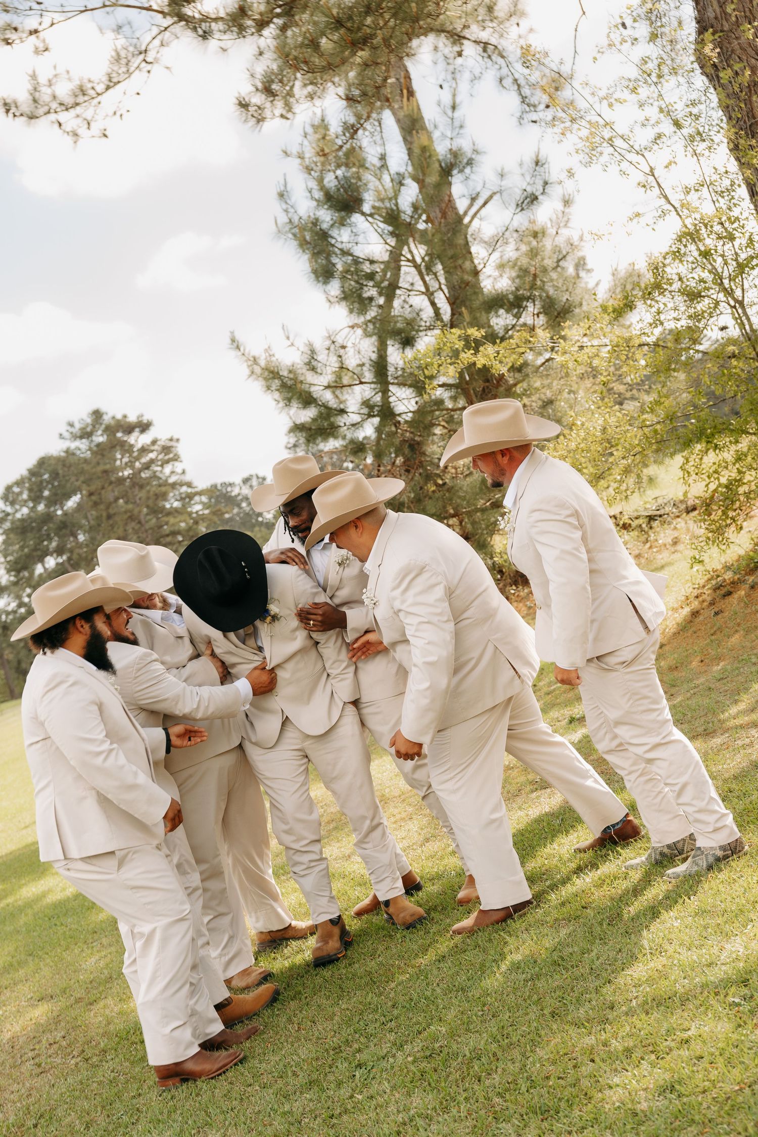 Groomsmen in matching cream suits and cowboy hats share a fun candid moment together on a grassy lawn.