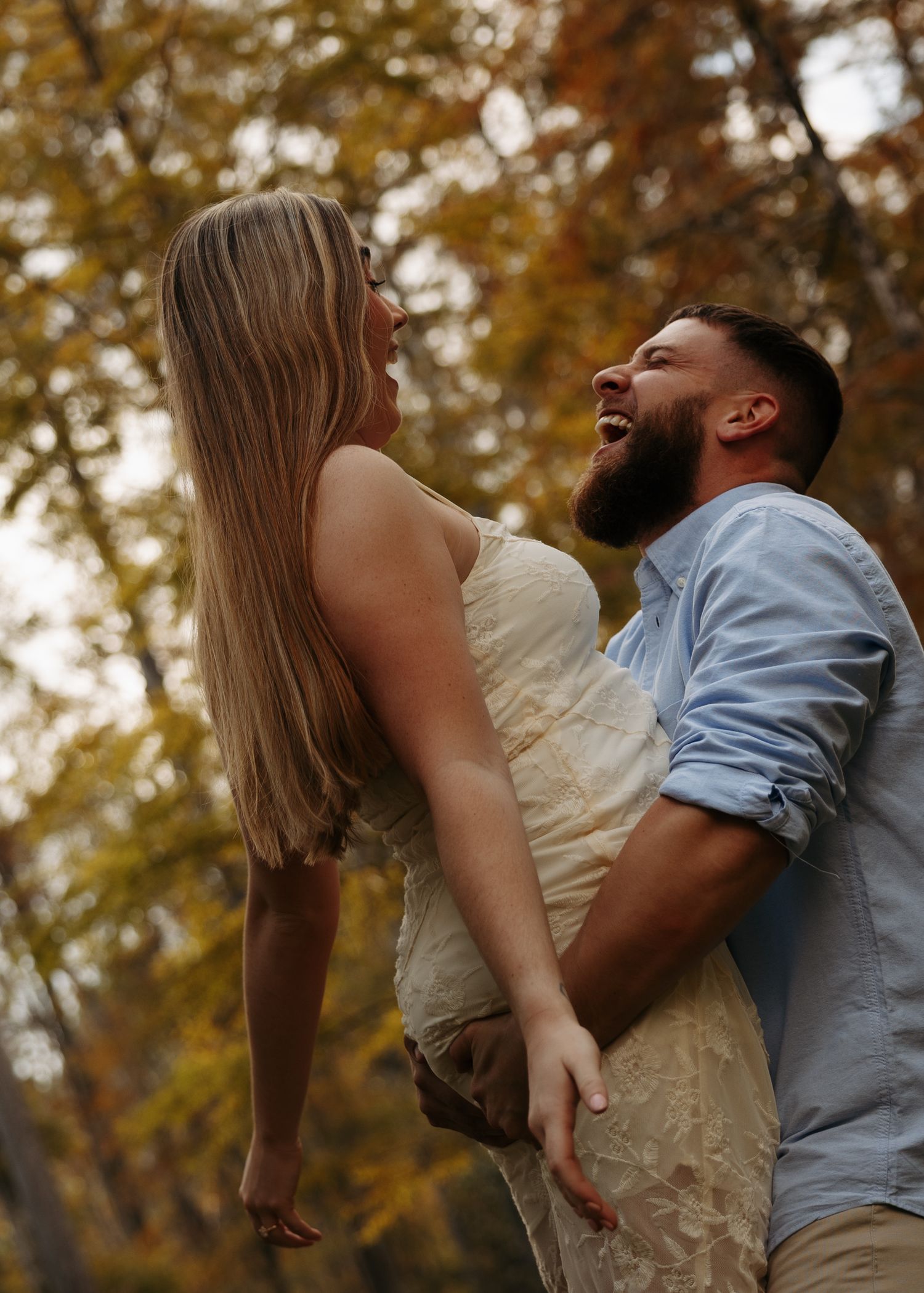 A bearded man joyfully lifts a blonde woman in a cream dress among autumn trees.
