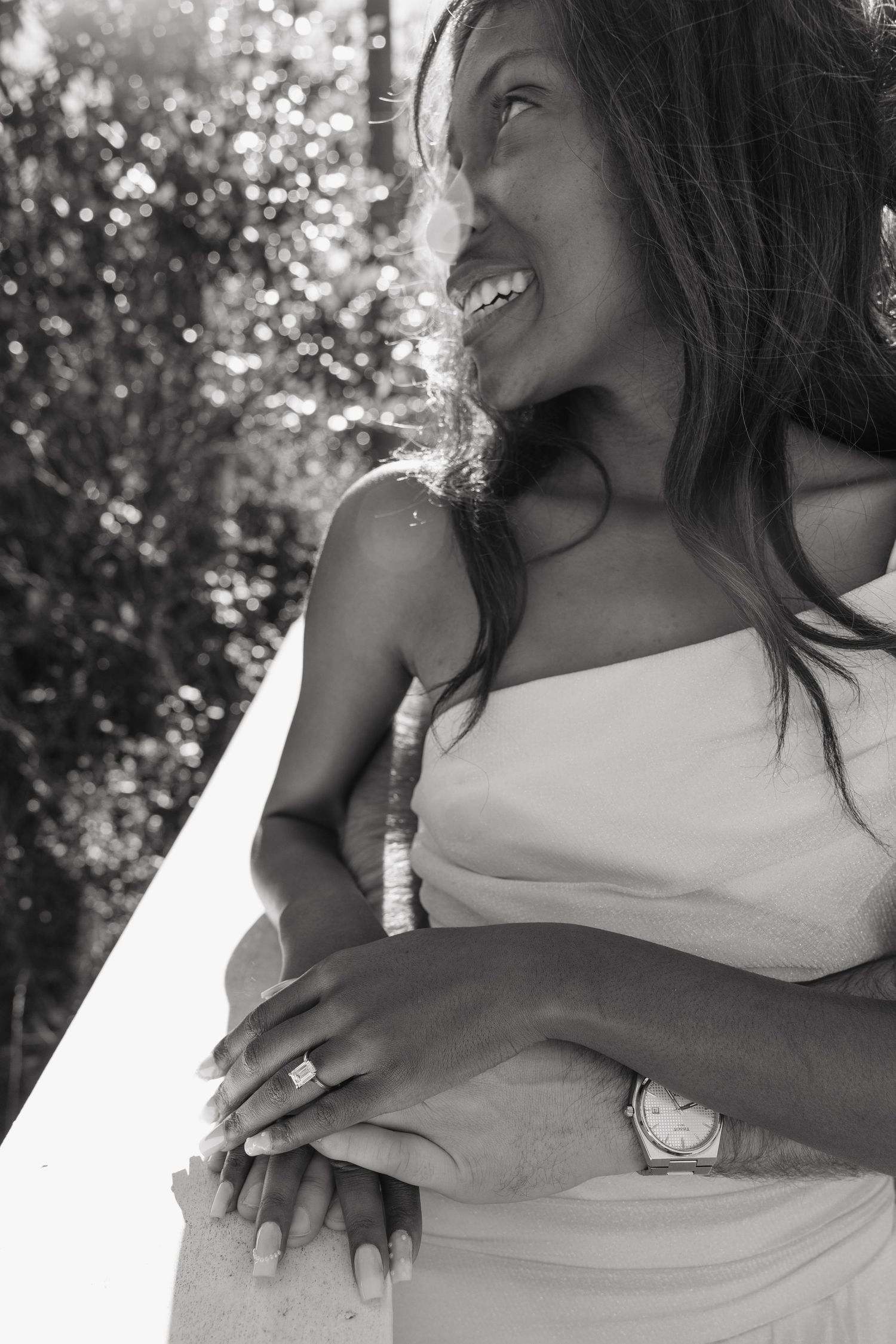 Black and white photo of a smiling bride in a strapless gown, showing her engagement ring and groom's watch.