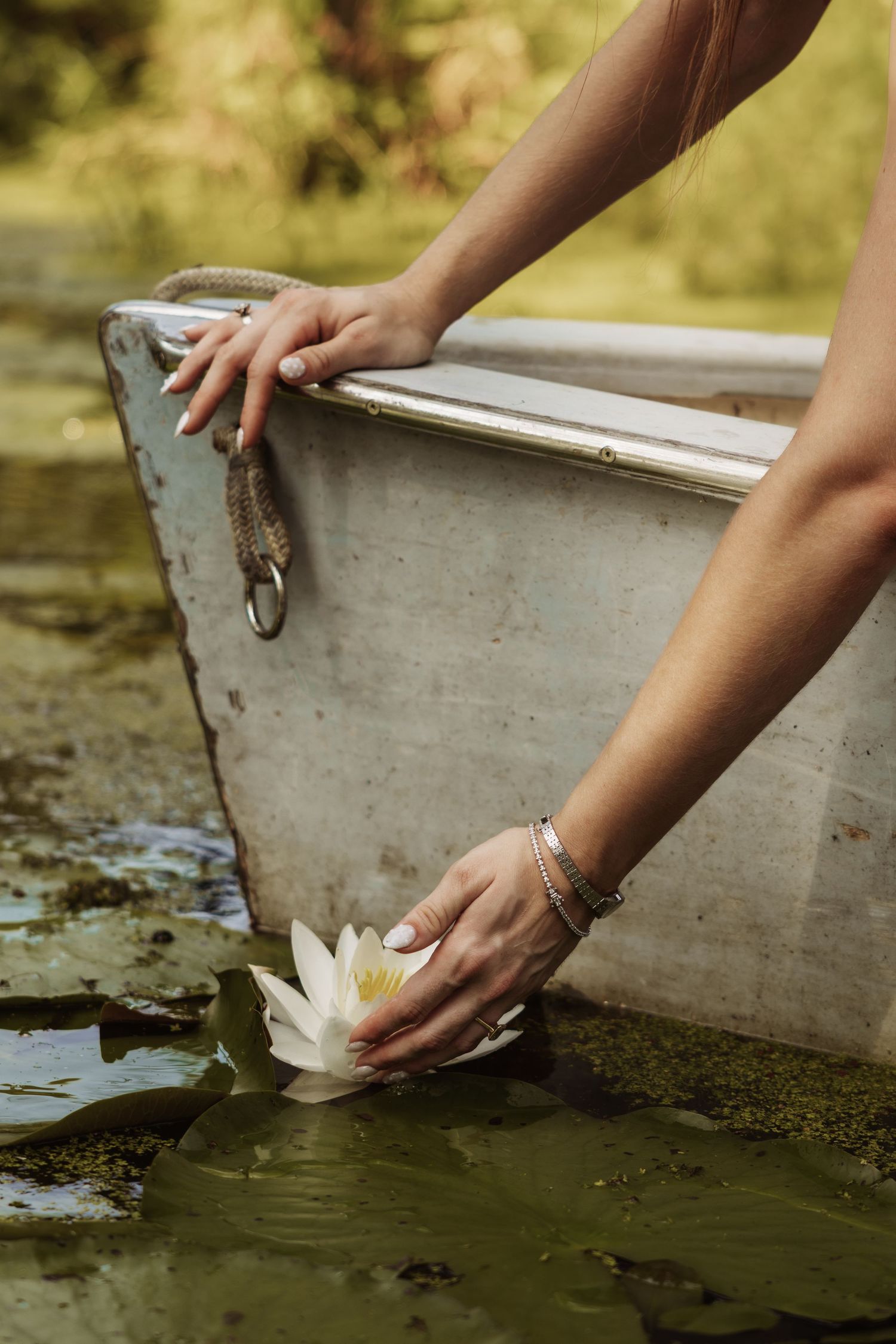 Woman reaching from a vintage white rowboat to touch a white water lily floating among lily pads on a calm pond.