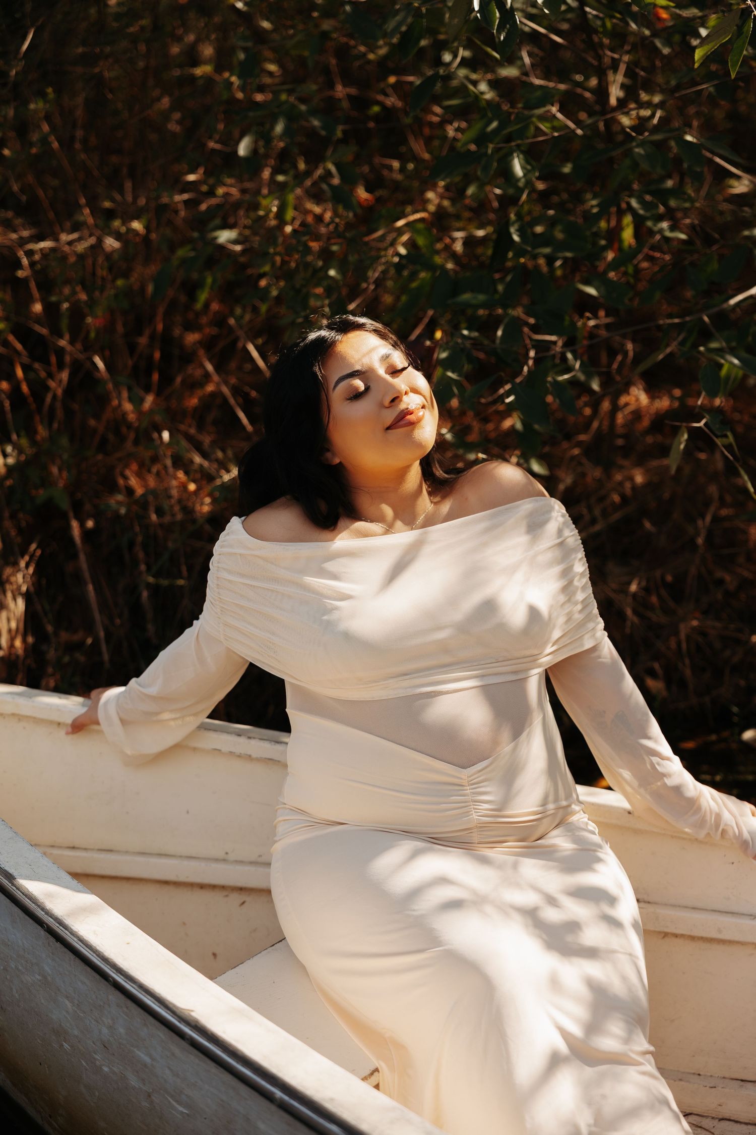 Woman in white off-shoulder dress sitting gracefully in a boat, bathed in warm sunlight with lush greenery behind her.