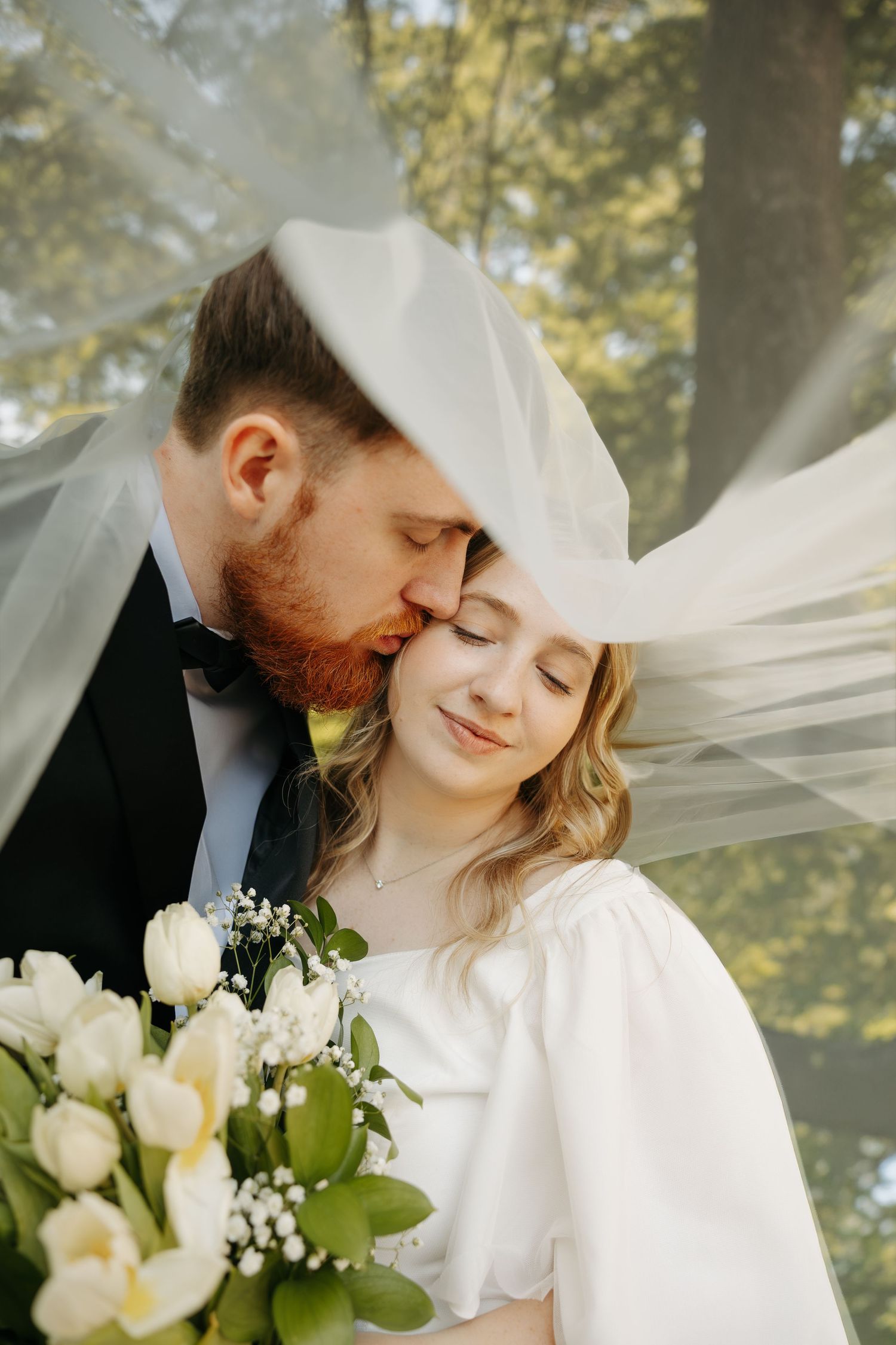 Groom kisses brides forehead under a flowing veil as she holds white tulips in a garden setting.