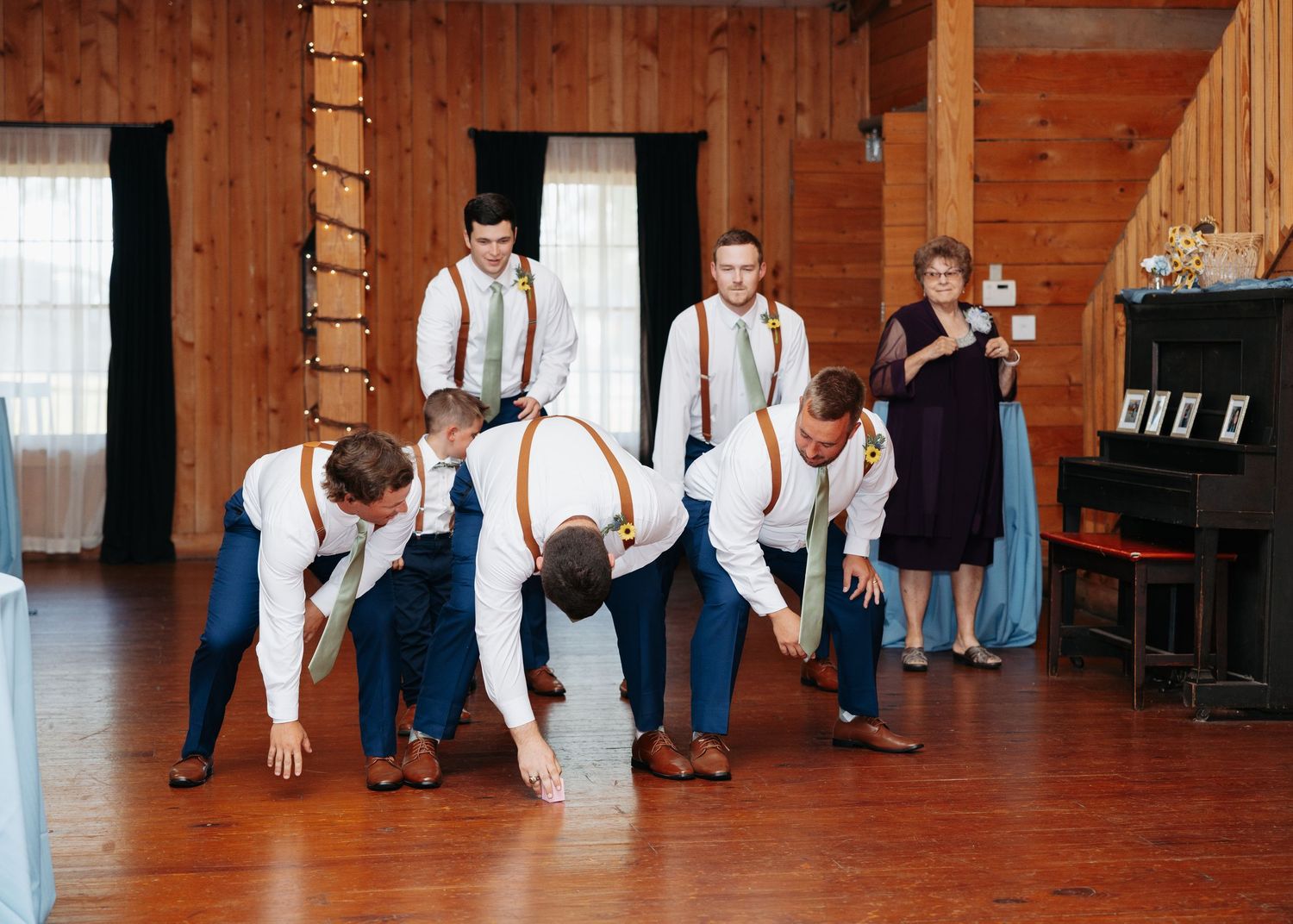 Groomsmen in white shirts and suspenders perform playful dance moves at wedding reception.