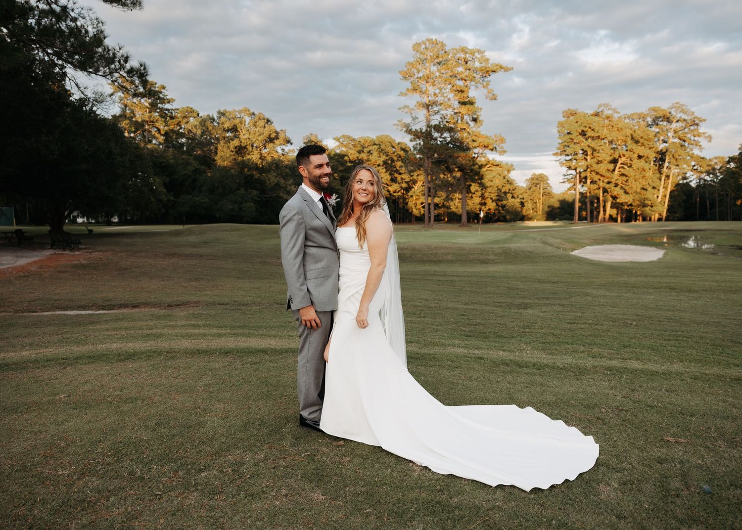 Bride and groom posing on a lush golf course at golden hour, her cathedral veil trailing behind.