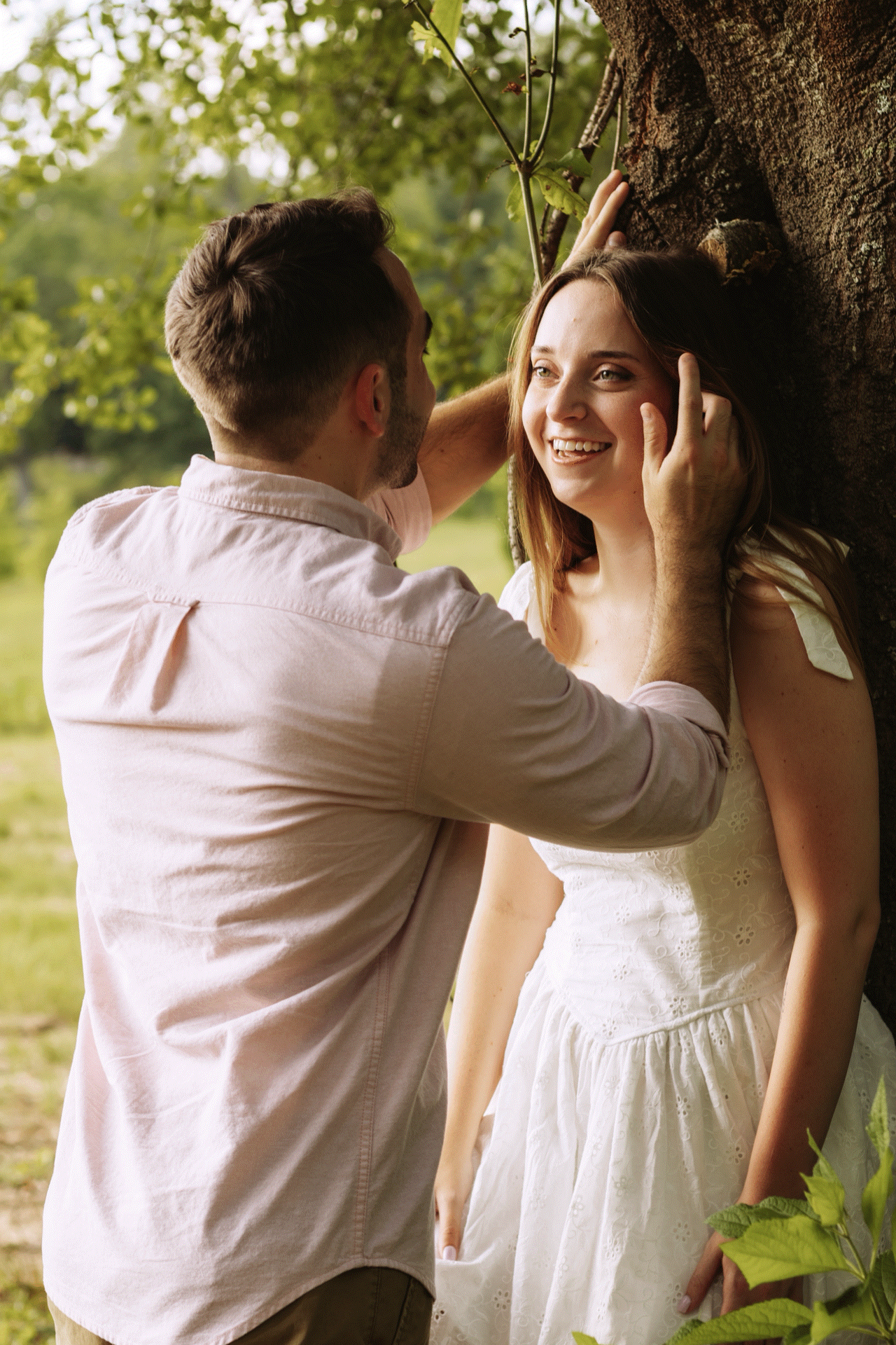 A smiling woman in a white dress leans against a tree as a man gently touches her face outdoors.