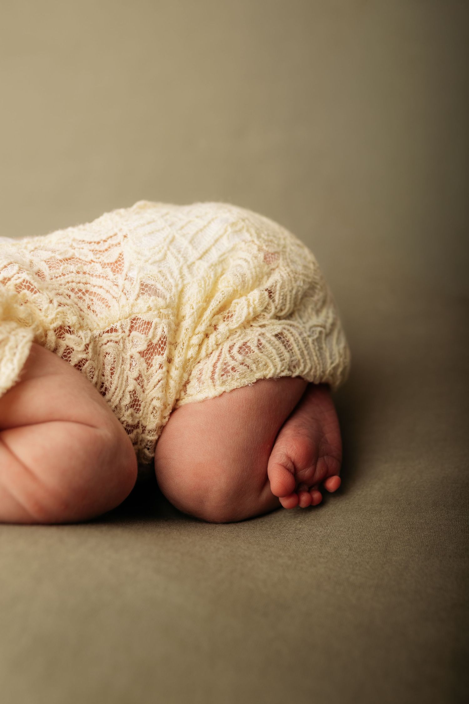 Newborn baby feet and legs in cream lace outfit on olive green backdrop, close-up photography.