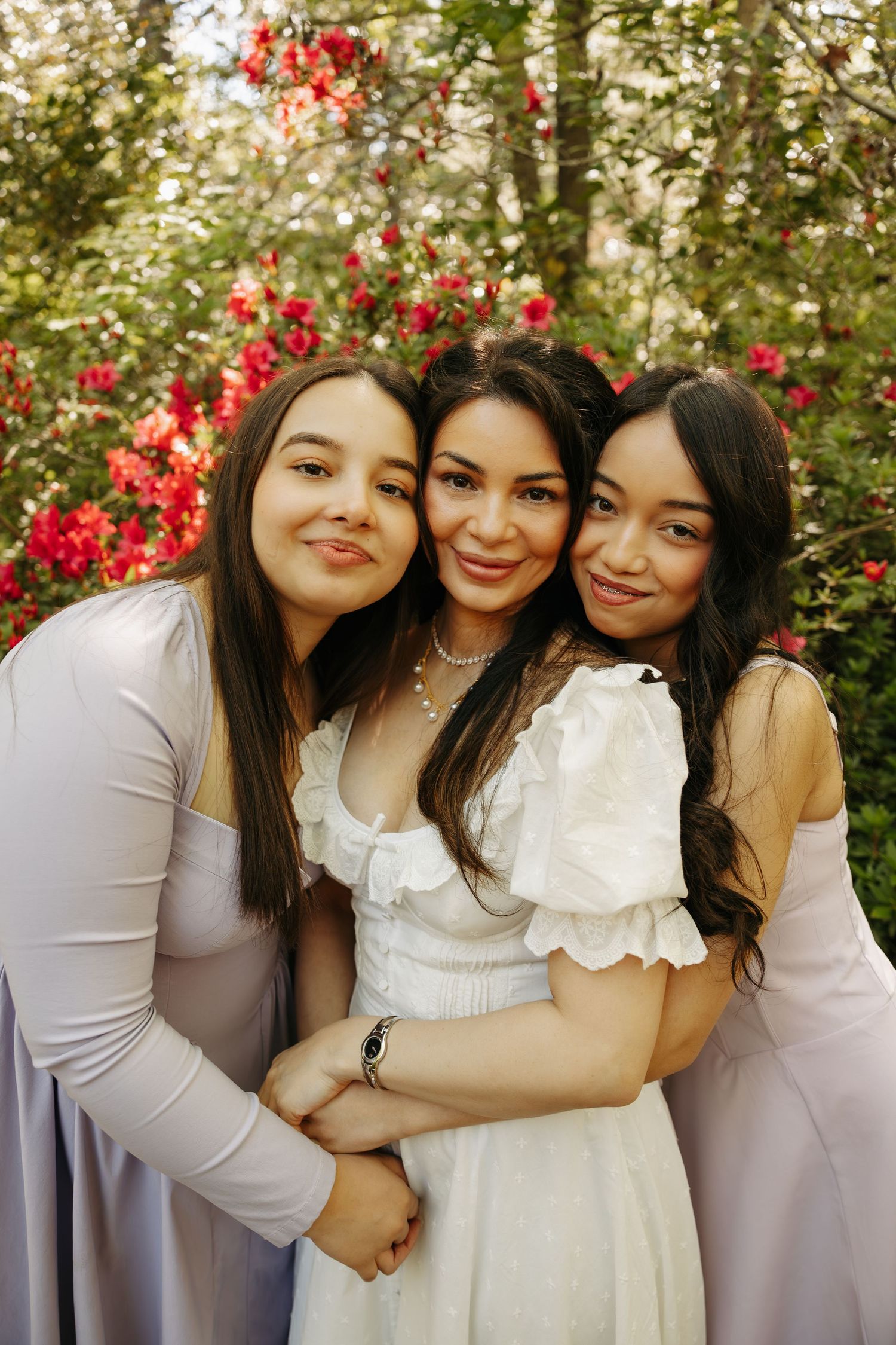 Three women in elegant dresses pose together outdoors, smiling warmly in front of vibrant red flowering bushes.