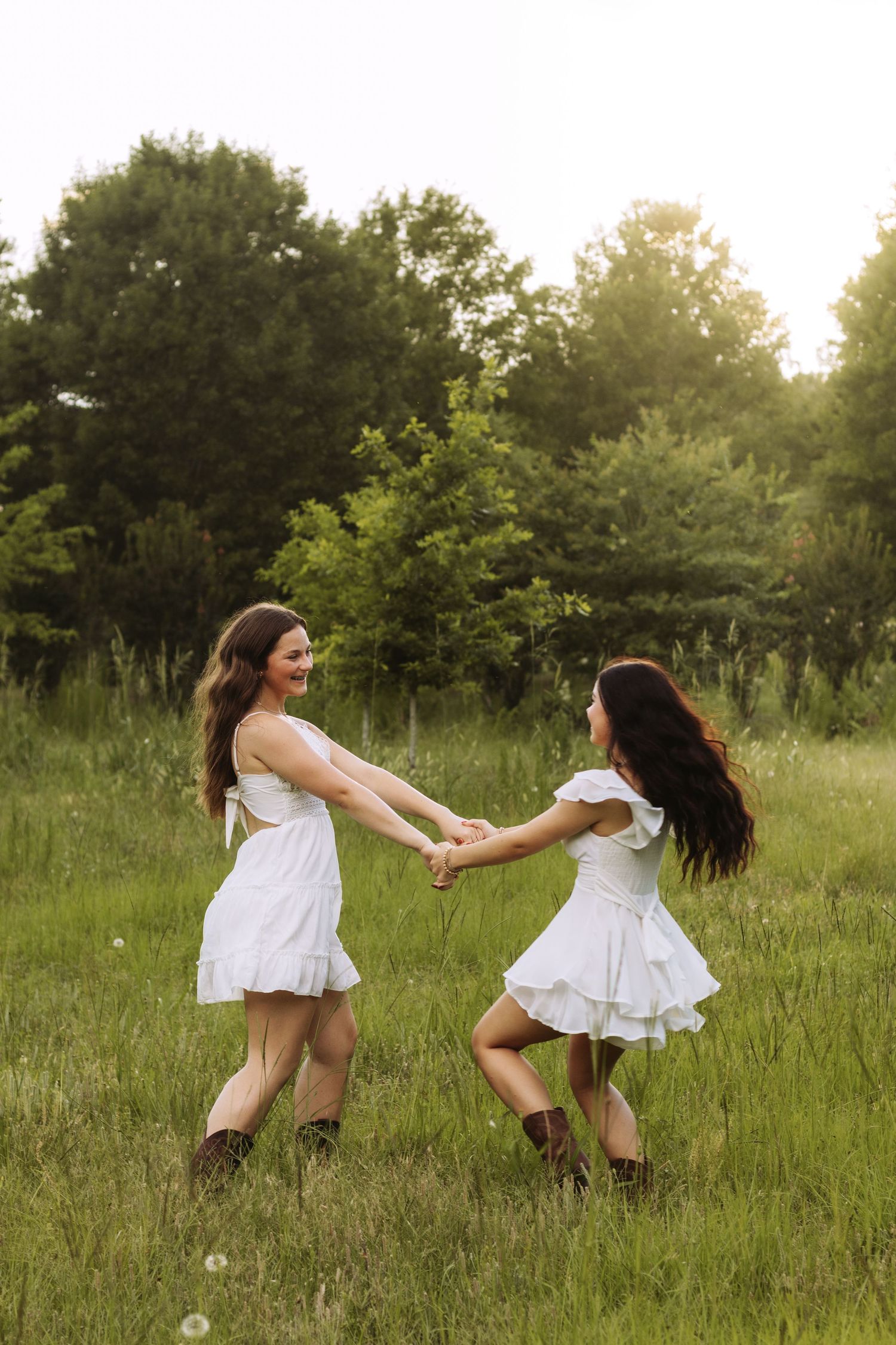 Two people in white dresses holding hands while dancing in a grassy meadow at sunset.