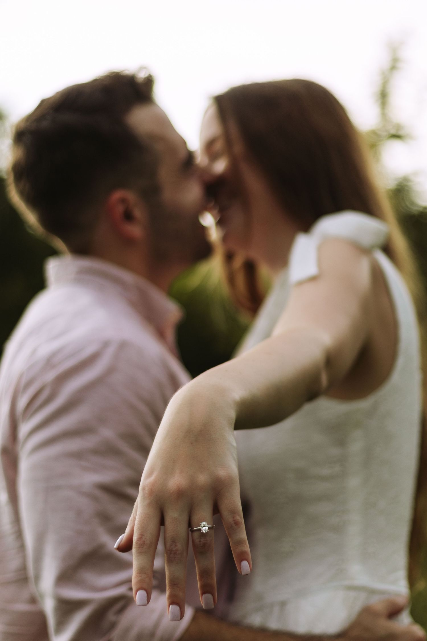 An engaged couple share an intimate moment as she displays her engagement ring in natural lighting.