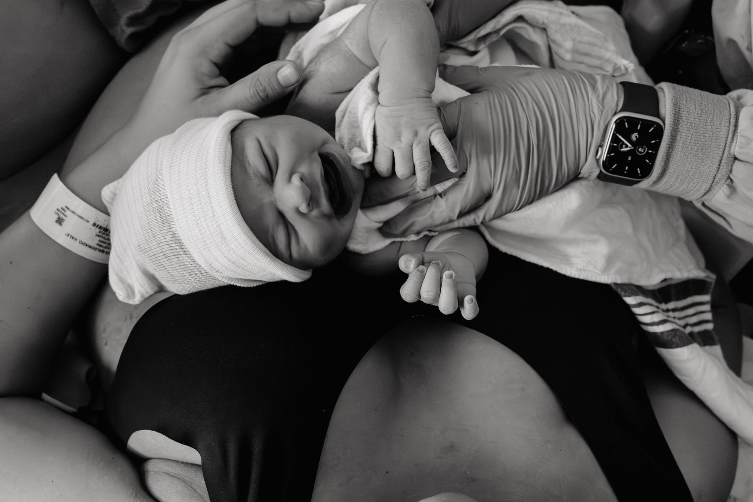 A baby lying on someone's lap stretches and yawns while wearing a white onesie, captured in black and white.