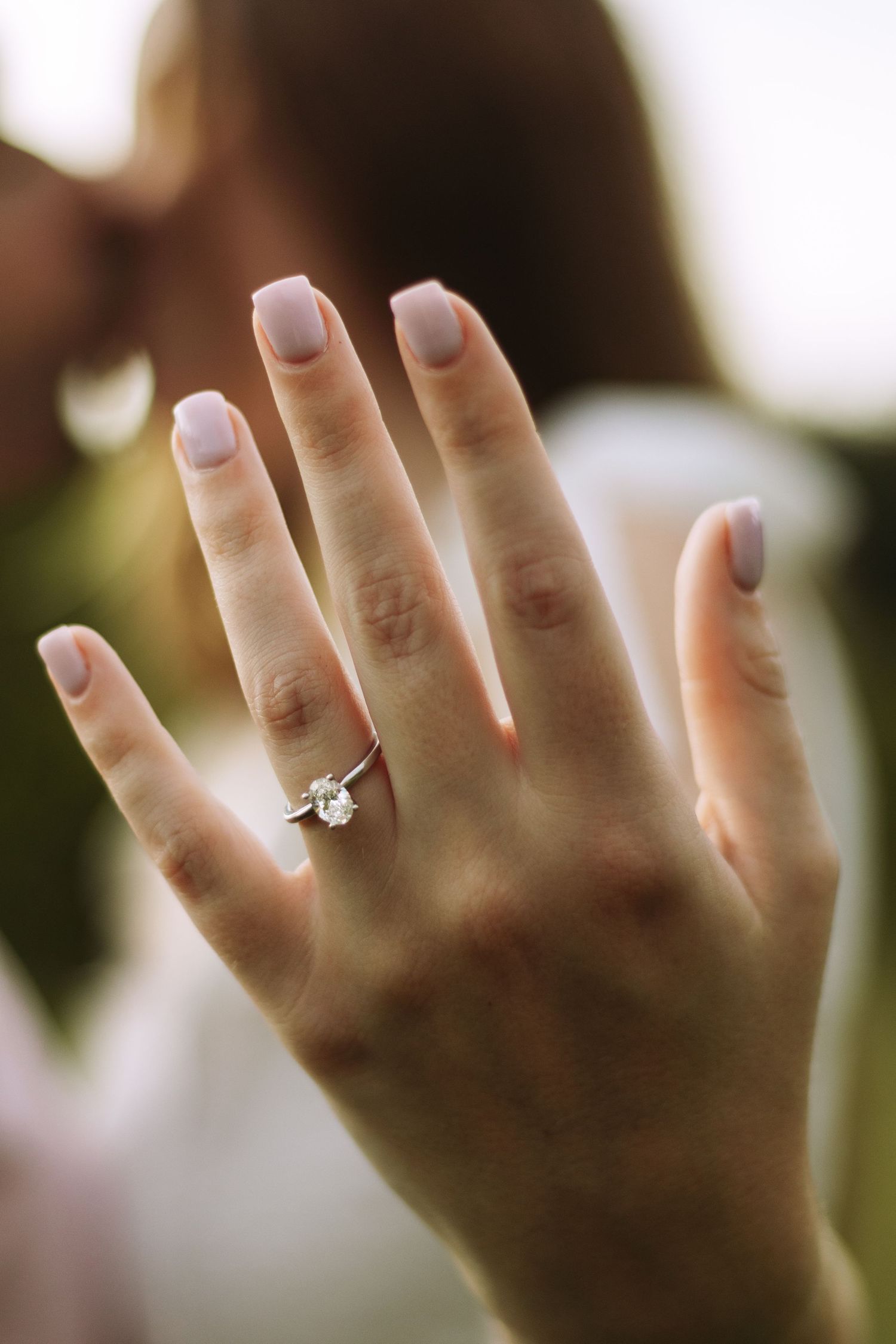 A hand wearing a sparkling diamond engagement ring shows off fresh pink manicured nails against a soft blurred background.