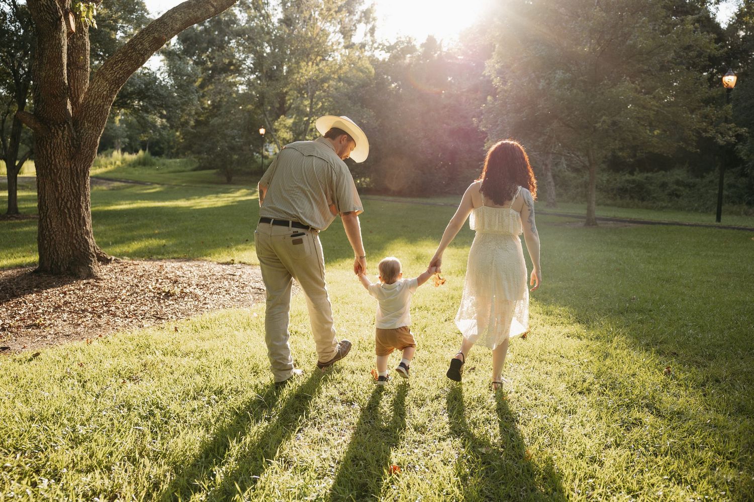 A couple holding hands walking in a sun-drenched park during golden hour as their long shadows stretch across green grass.