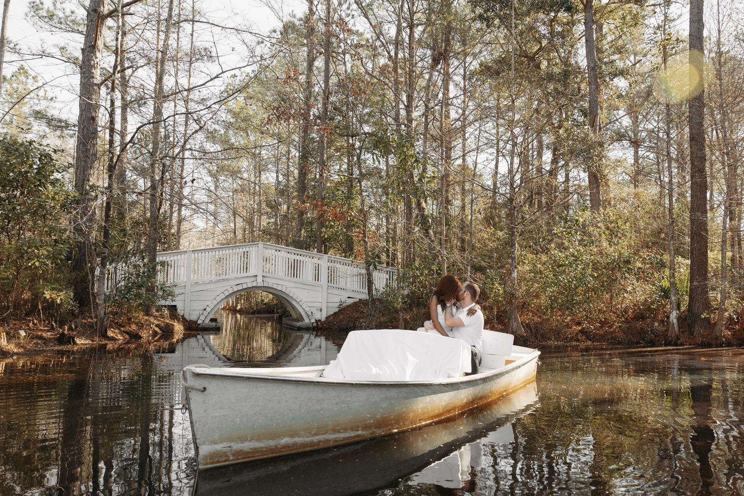 Bride and groom share a romantic kiss in a white boat on a serene lake surrounded by tall trees.