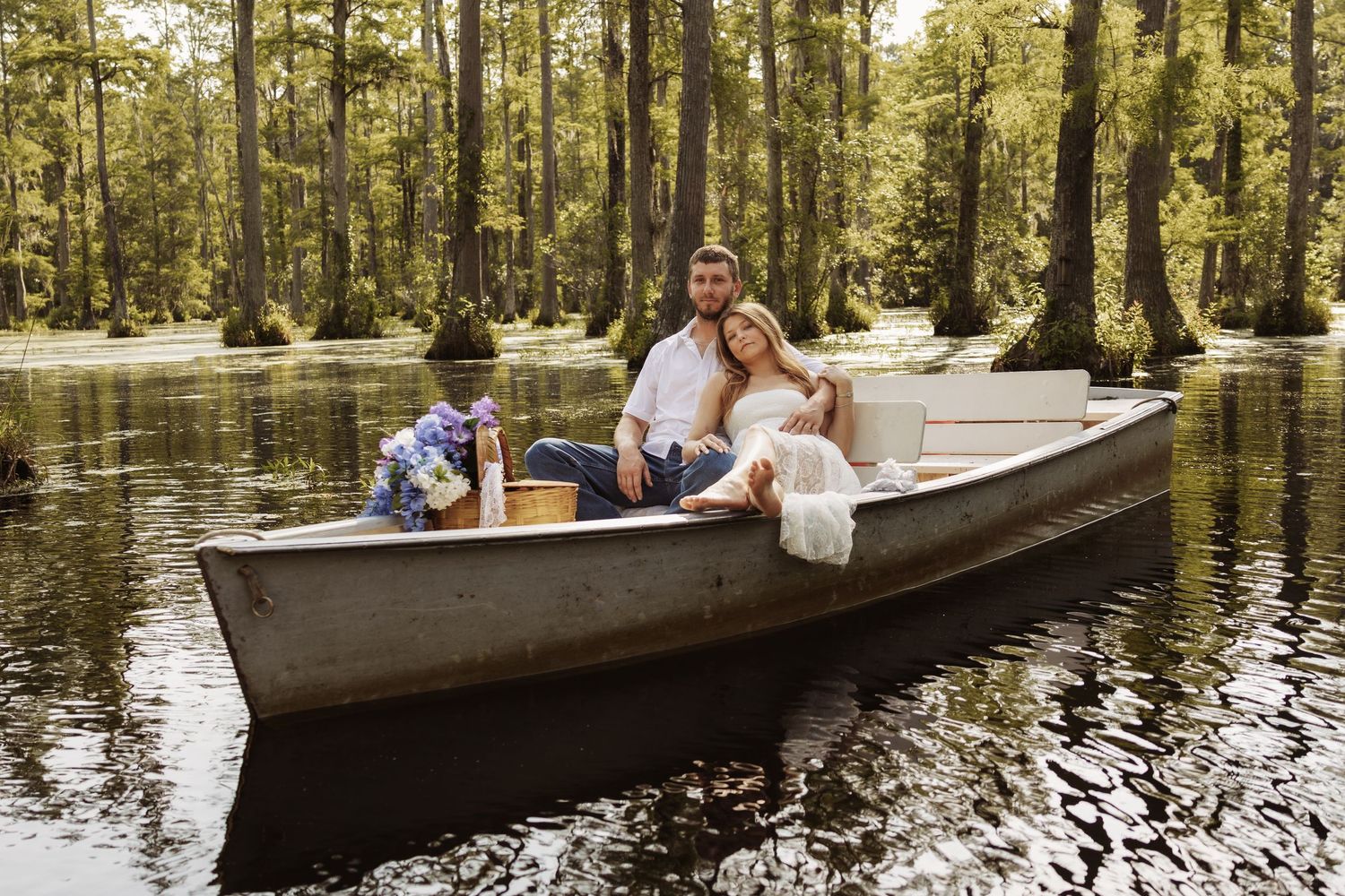 Couple in wedding attire relaxing in a canoe on a serene swamp surrounded by cypress trees.
