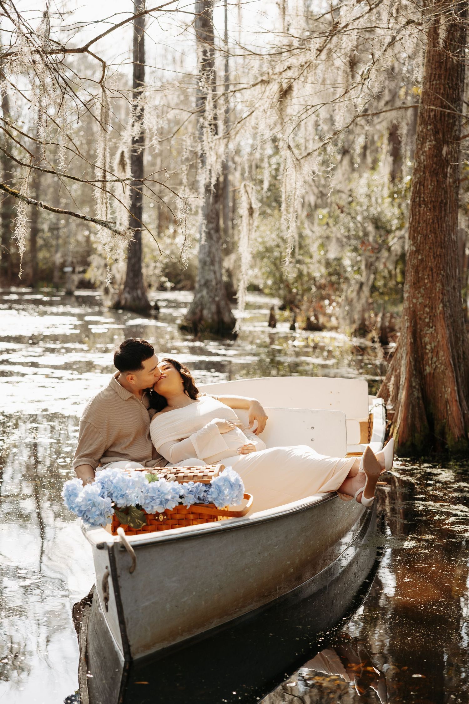 Couple sharing a romantic kiss in a white boat surrounded by cypress trees and blue hydrangeas in a serene swamp.