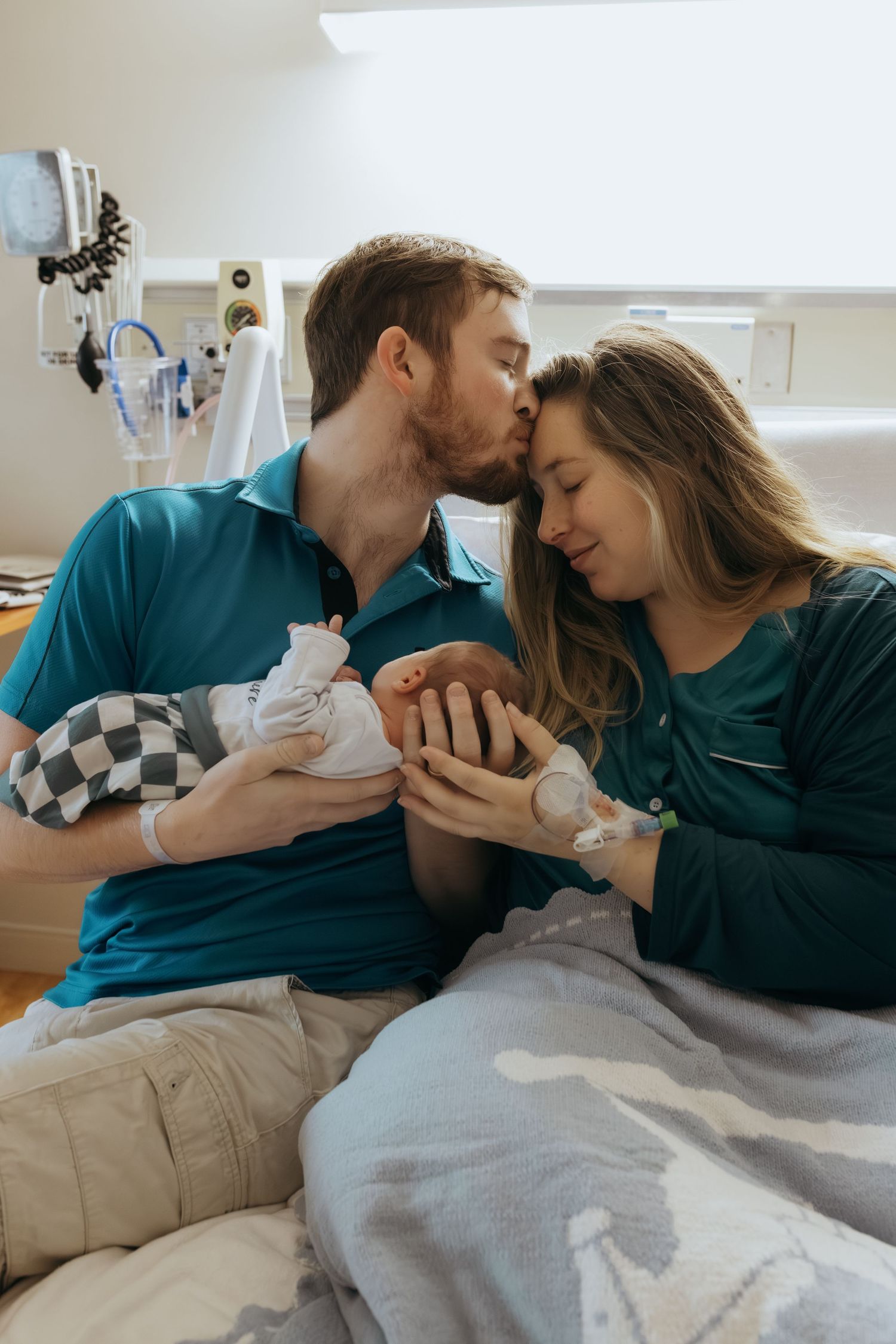 New parents share a tender kiss while holding their newborn baby in a hospital room after delivery.