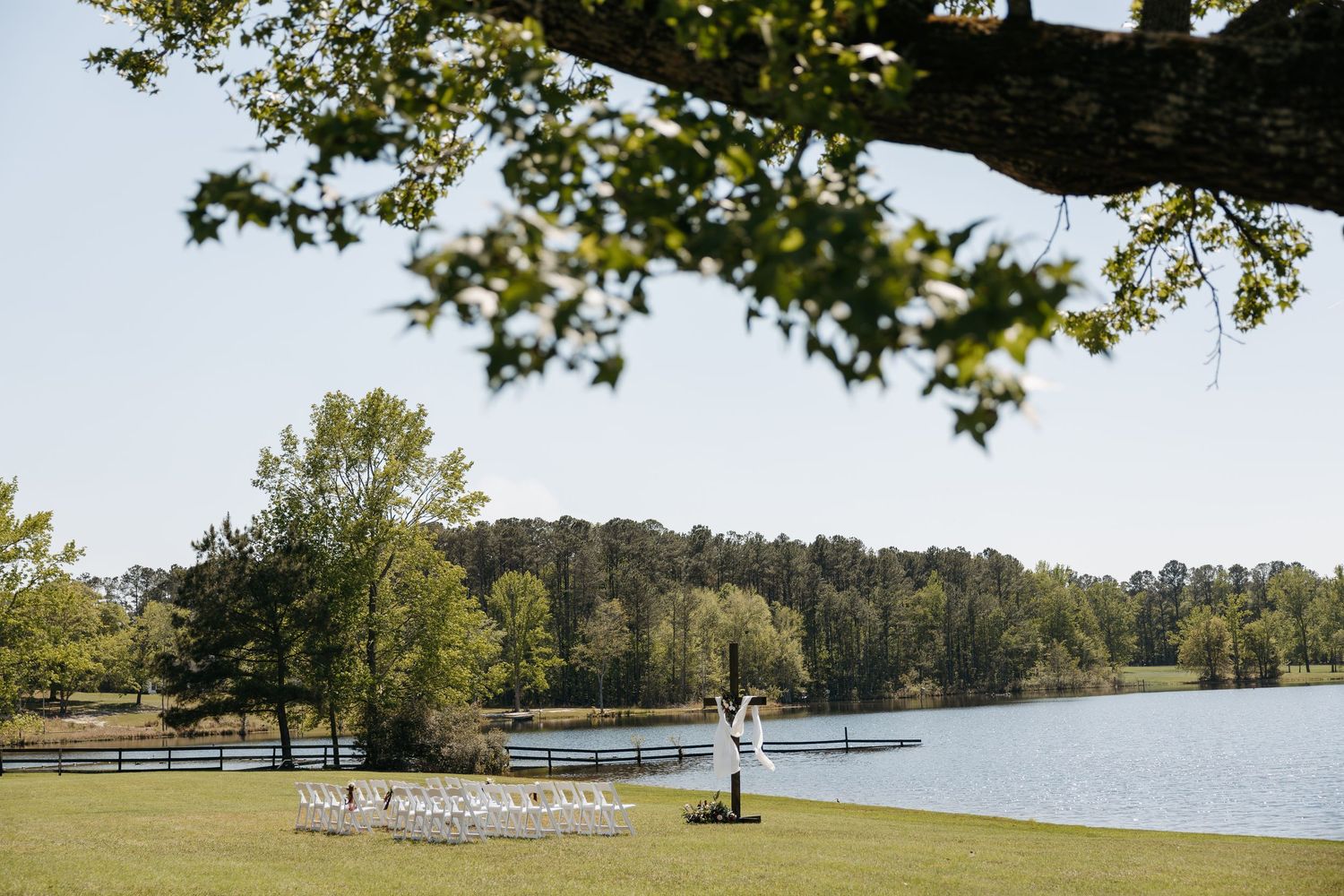 A bride and groom pose by a serene lakeside venue surrounded by lush green trees and manicured lawns on a clear day.