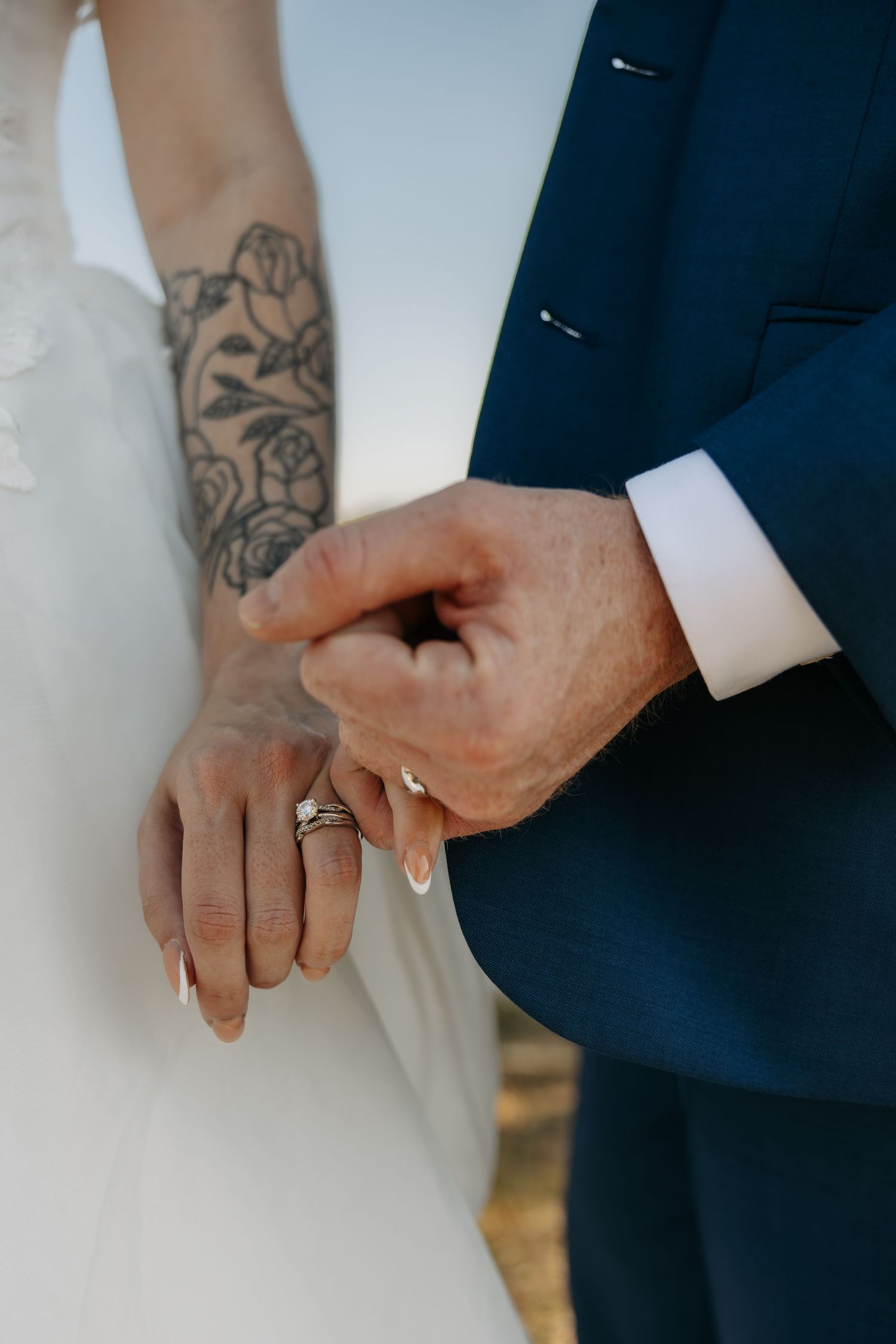 Close-up of bride and groom hands showing wedding rings, bride has rose tattoo on forearm, groom wears navy suit.