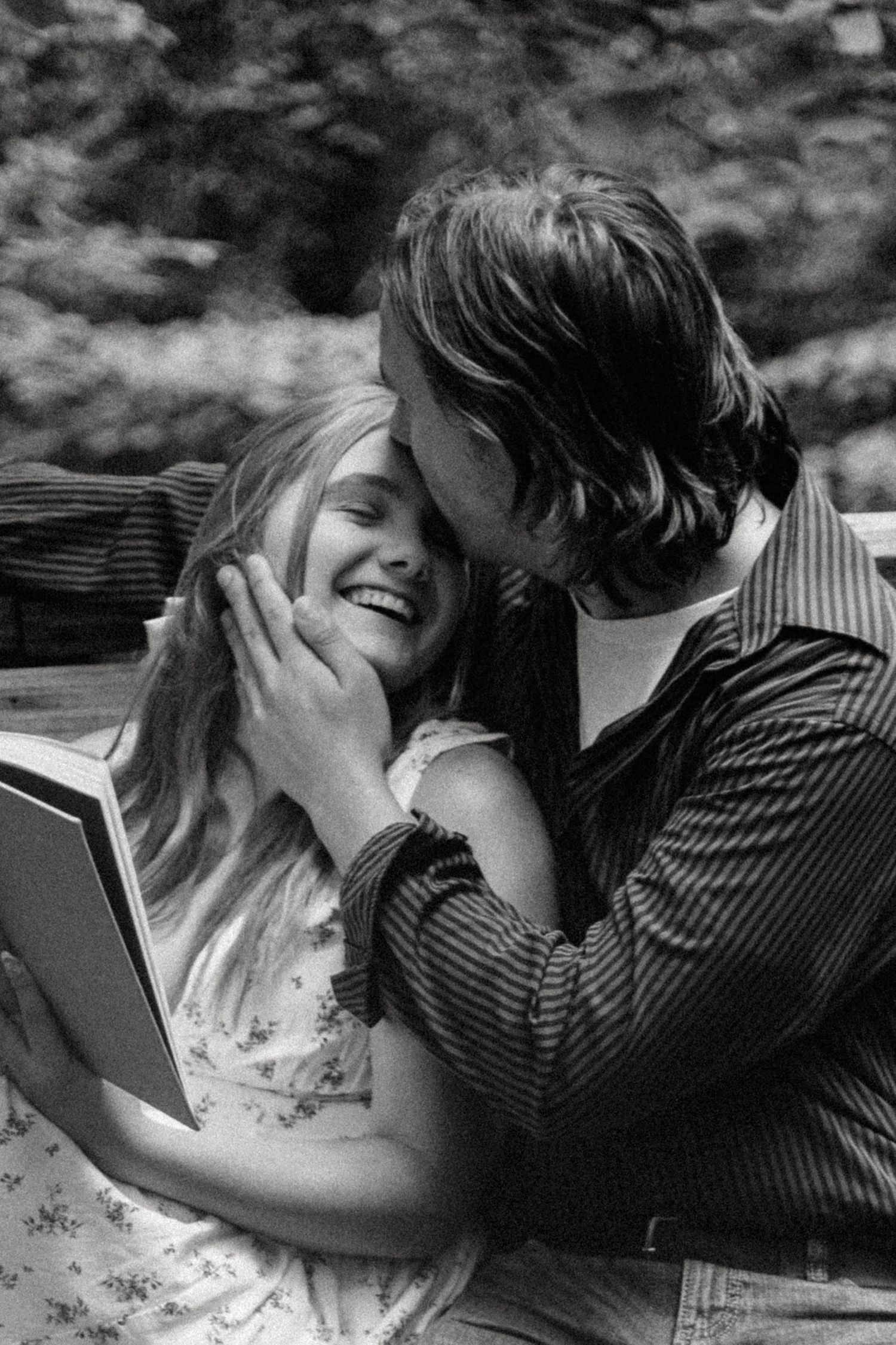 Black and white photo of a smiling woman holding a book while a man tenderly kisses her cheek outdoors.
