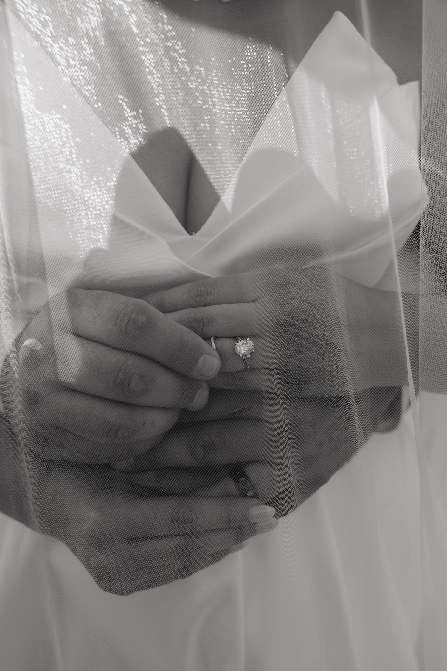 Black and white close-up of bride and grooms hands clasped together showing diamond engagement ring.