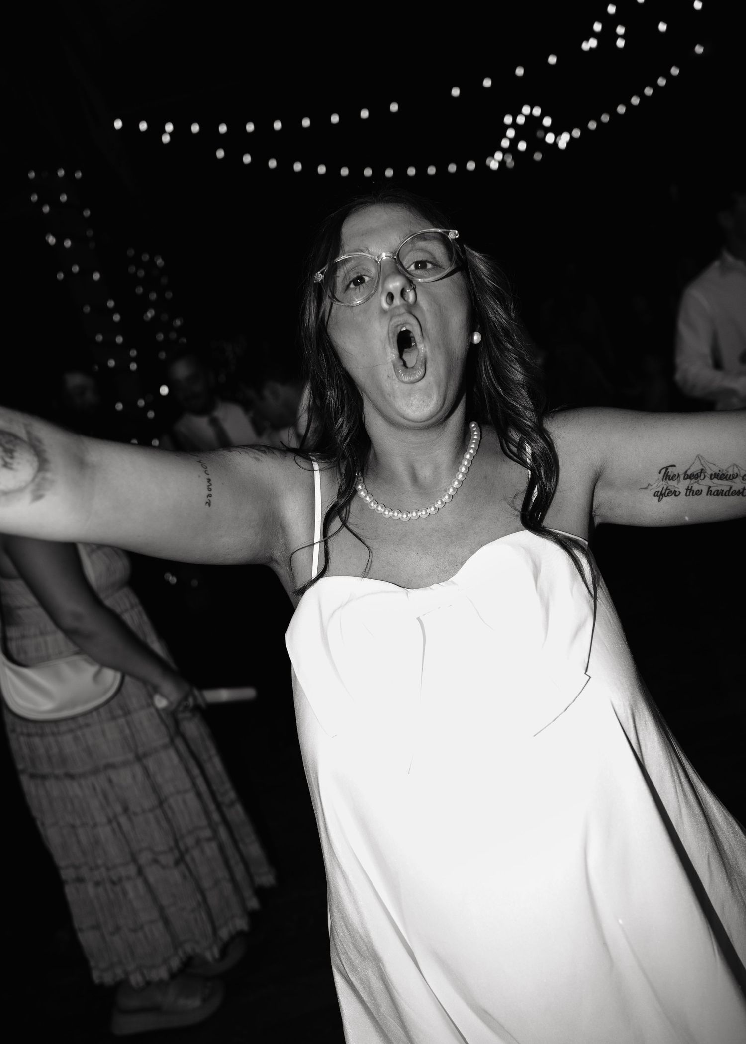 Bride in white dress and pearls dances joyfully with arms outstretched at wedding reception.