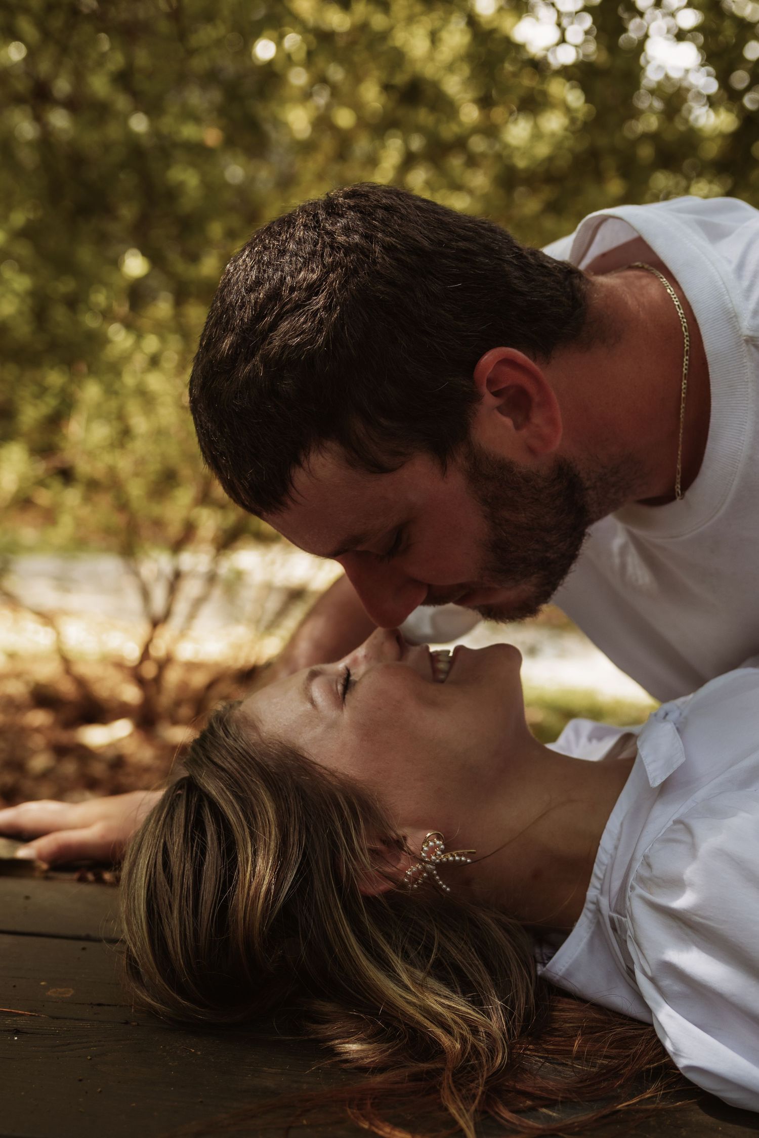A bearded man in white leans down to kiss a woman lying back on a wooden surface outdoors.