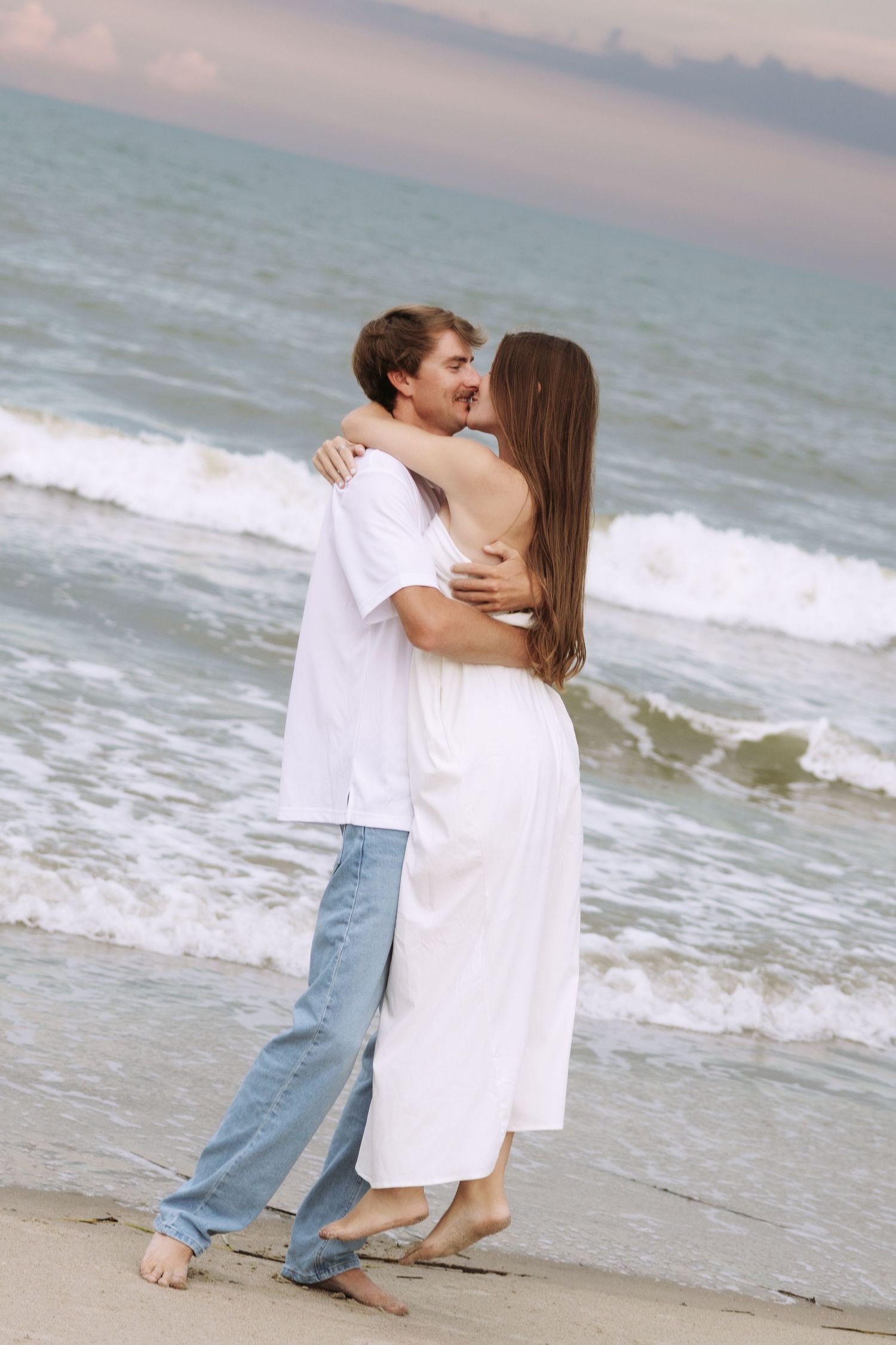 A romantic couple embraces and kisses on a sandy beach at sunset, wearing white and blue casual outfits.