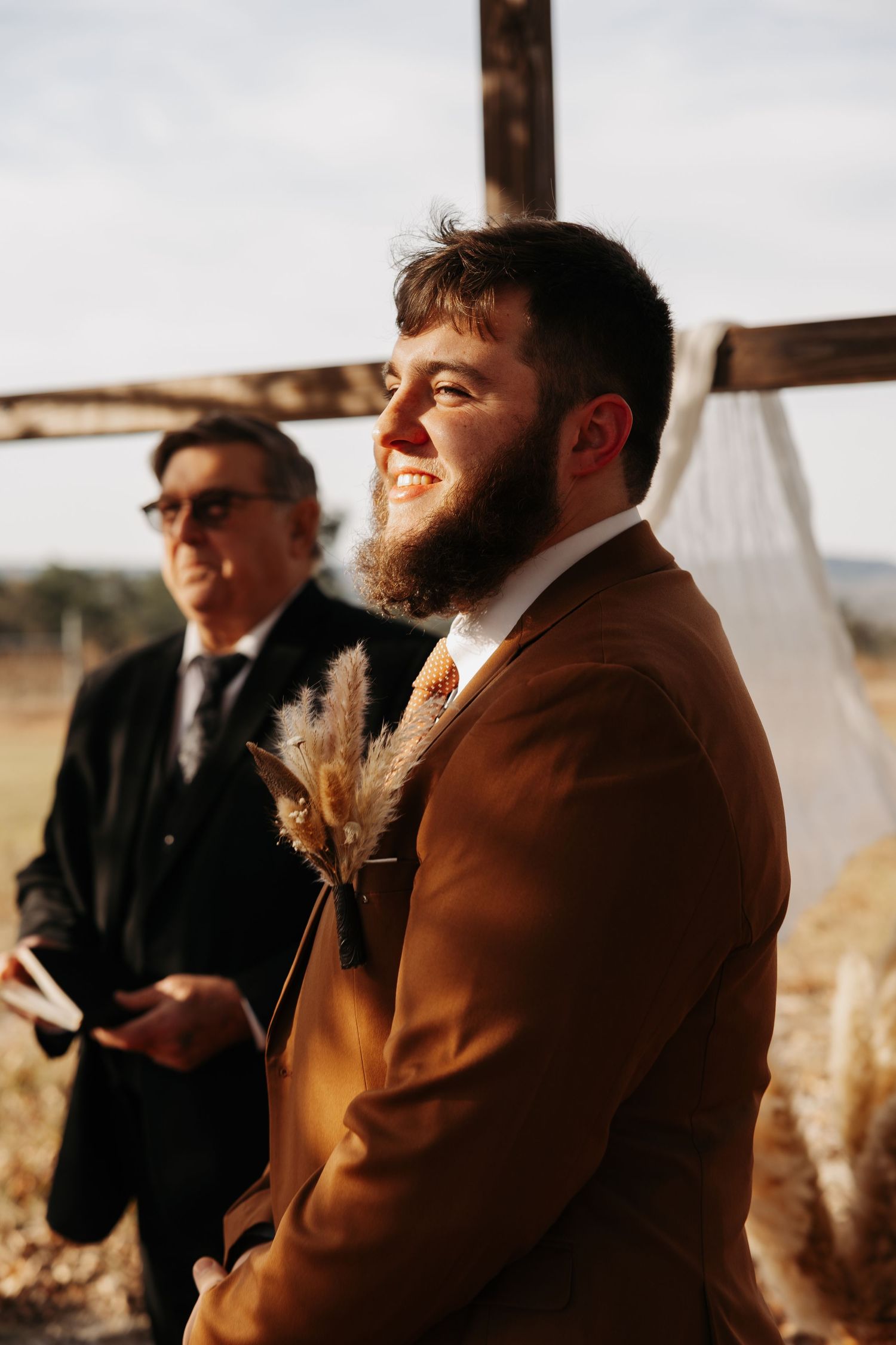 Smiling bearded groom in brown suit with pampas grass boutonniere awaits bride at the altar.