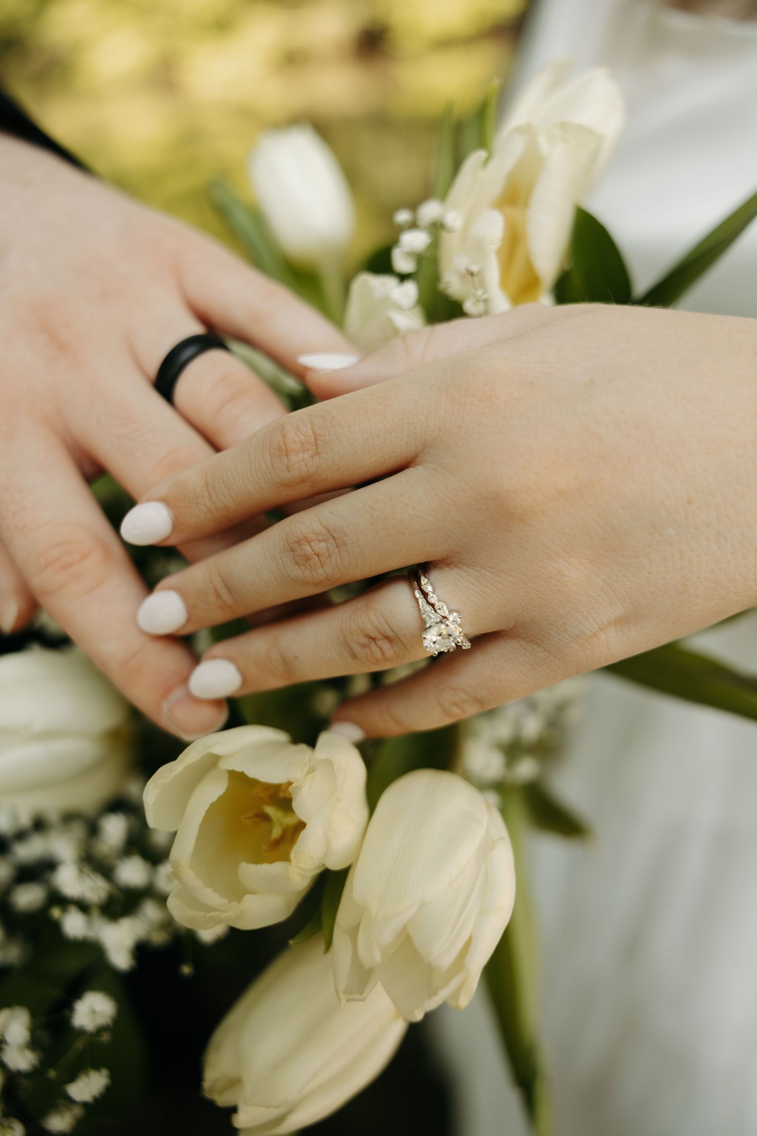 Newlyweds display wedding rings over a bouquet of white tulips, bride wearing a diamond ring.