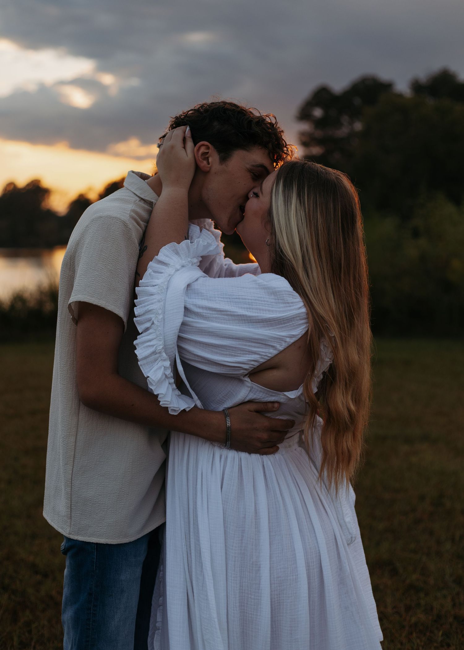 Couple sharing a romantic kiss at sunset in a field, woman in white ruffled dress, man in beige shirt and jeans.