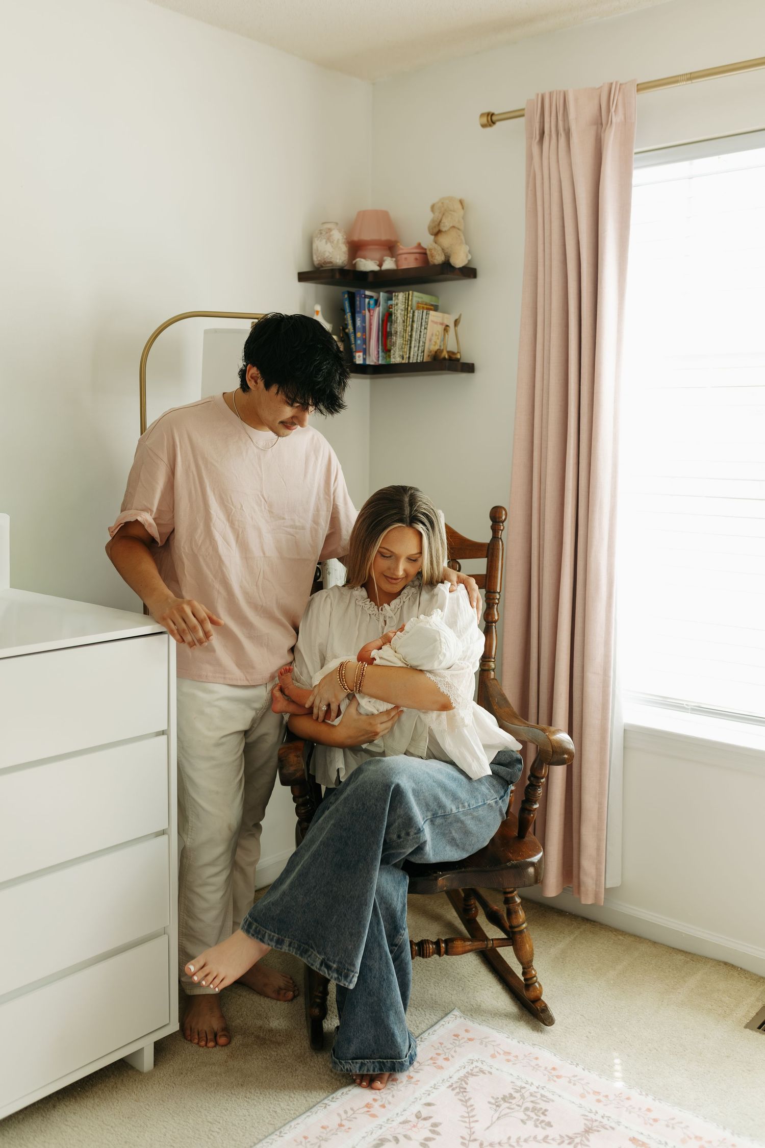 New parents admire their newborn baby in a soft pink nursery, mother seated in a wooden rocking chair.