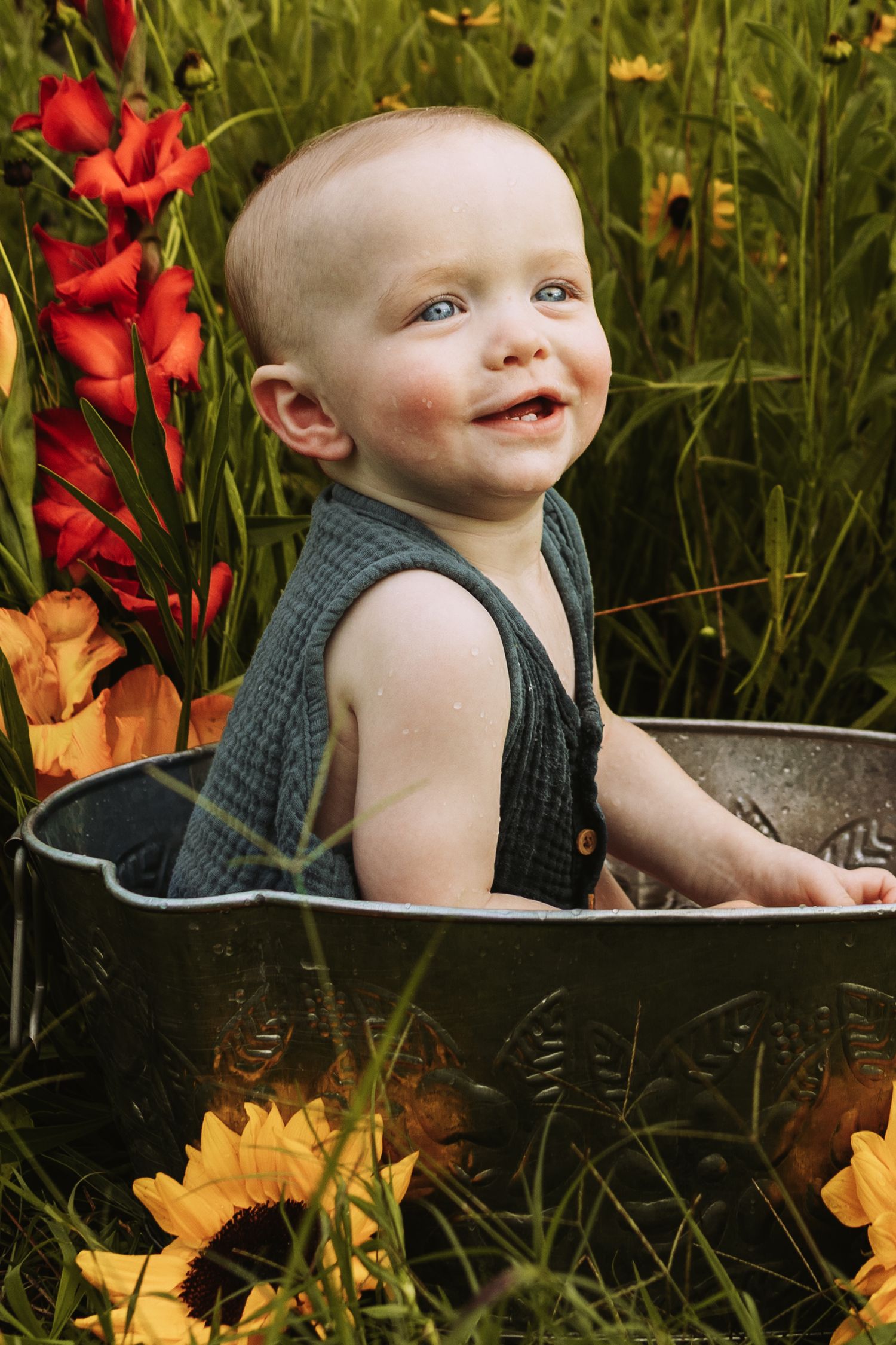 Small child sitting in vintage metal tub outdoors with red and yellow flowers in the background.