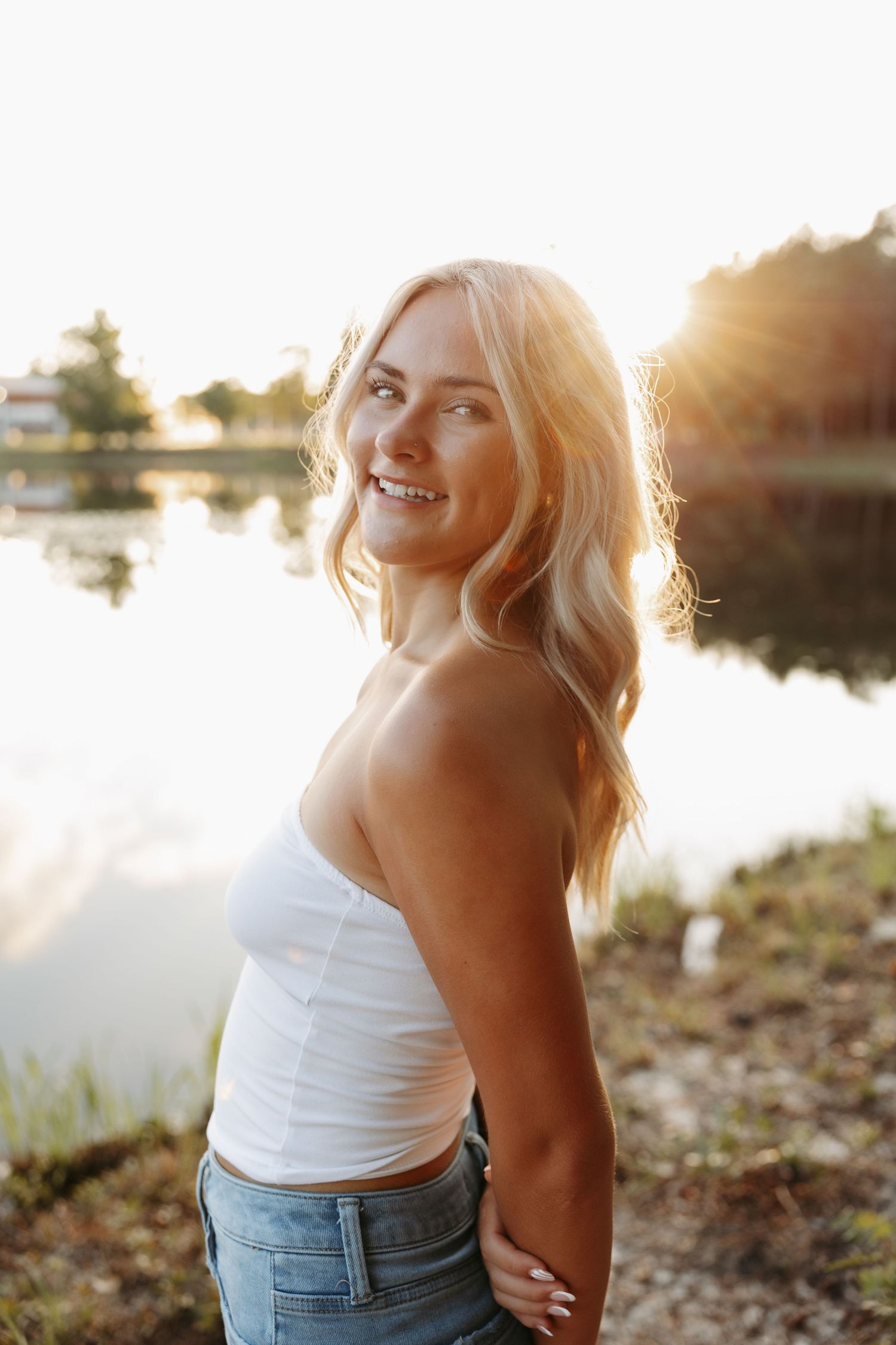 A person in a white tank top and jeans poses by a lake at sunset with golden light creating a backlit glow.