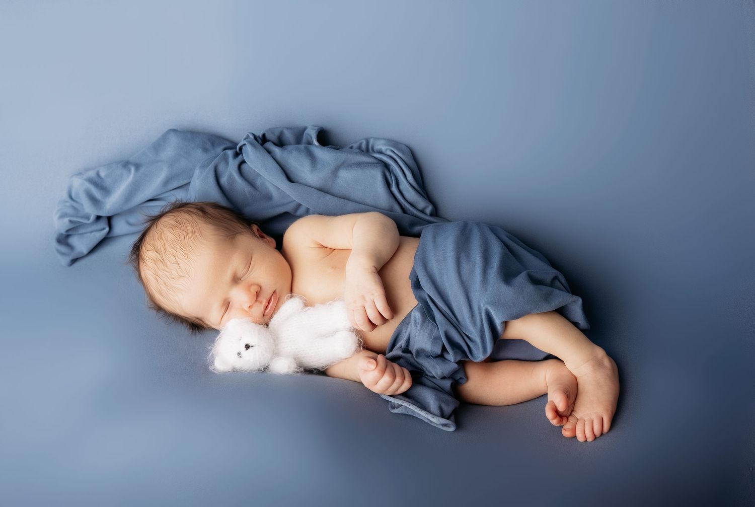 A sleeping baby lies peacefully on a blue-grey background in a minimalist newborn photography composition.