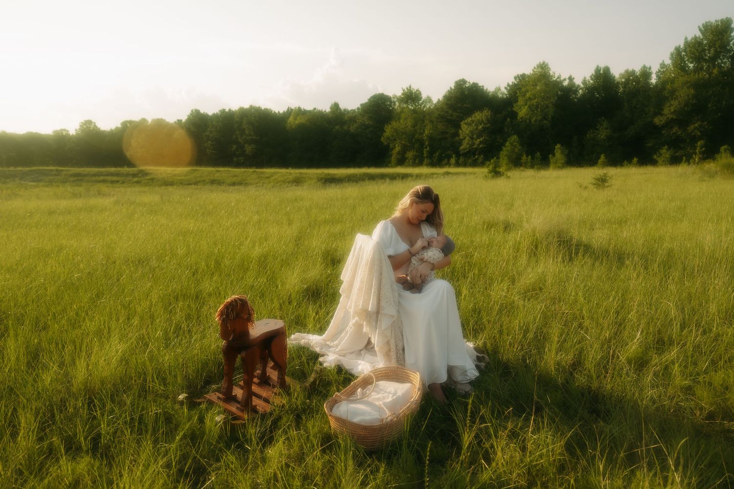 A woman in a white dress holds a baby in a sunlit green meadow with a wicker basket and rocking horse nearby.