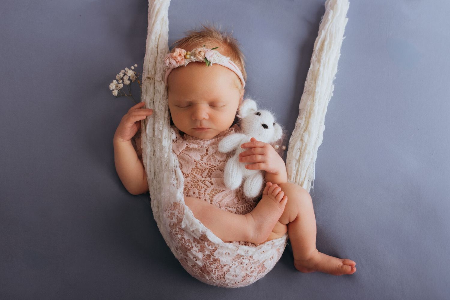 Newborn baby girl sleeping in lace wrap swing, holding white teddy bear, wearing pink floral headband.
